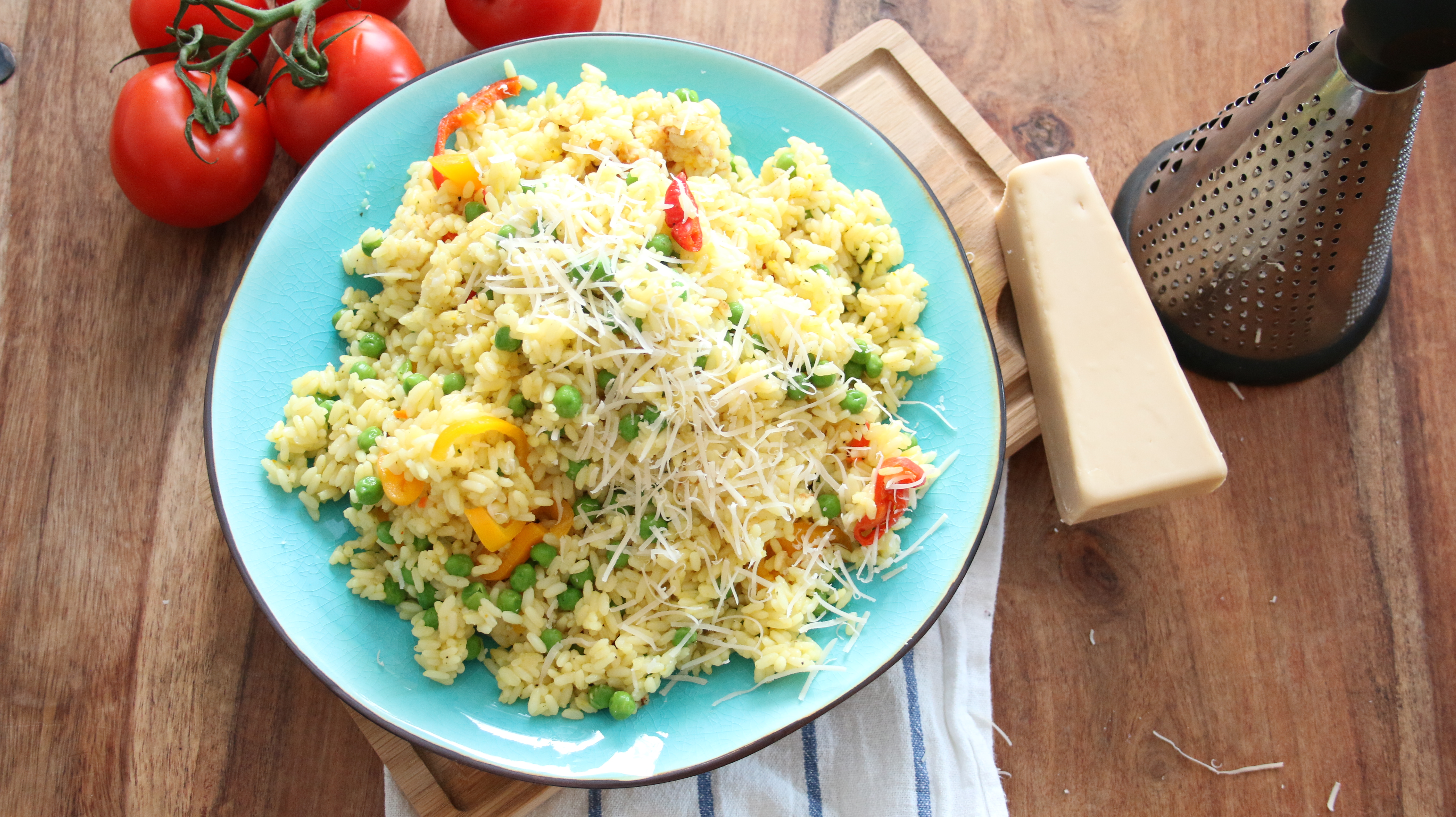 Risotto med gröna ärtor på en turkos tallrik, bredvid tallriken ligger en vegansk parmesanost och några tomater