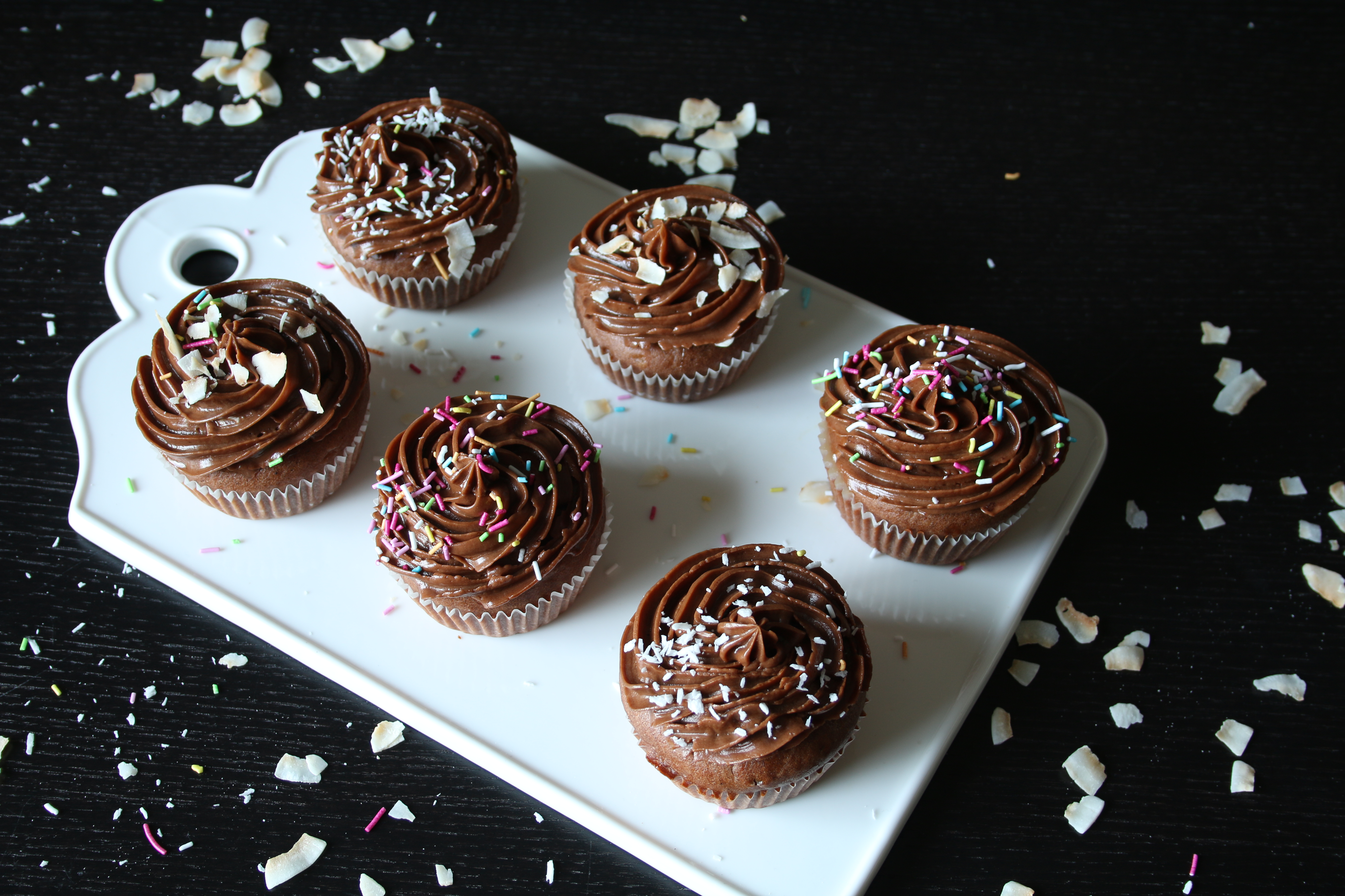 Sex stycken kolamuffins med mockaglasyr, kokos och strössel på en vit skärbräda på ett svart bord.