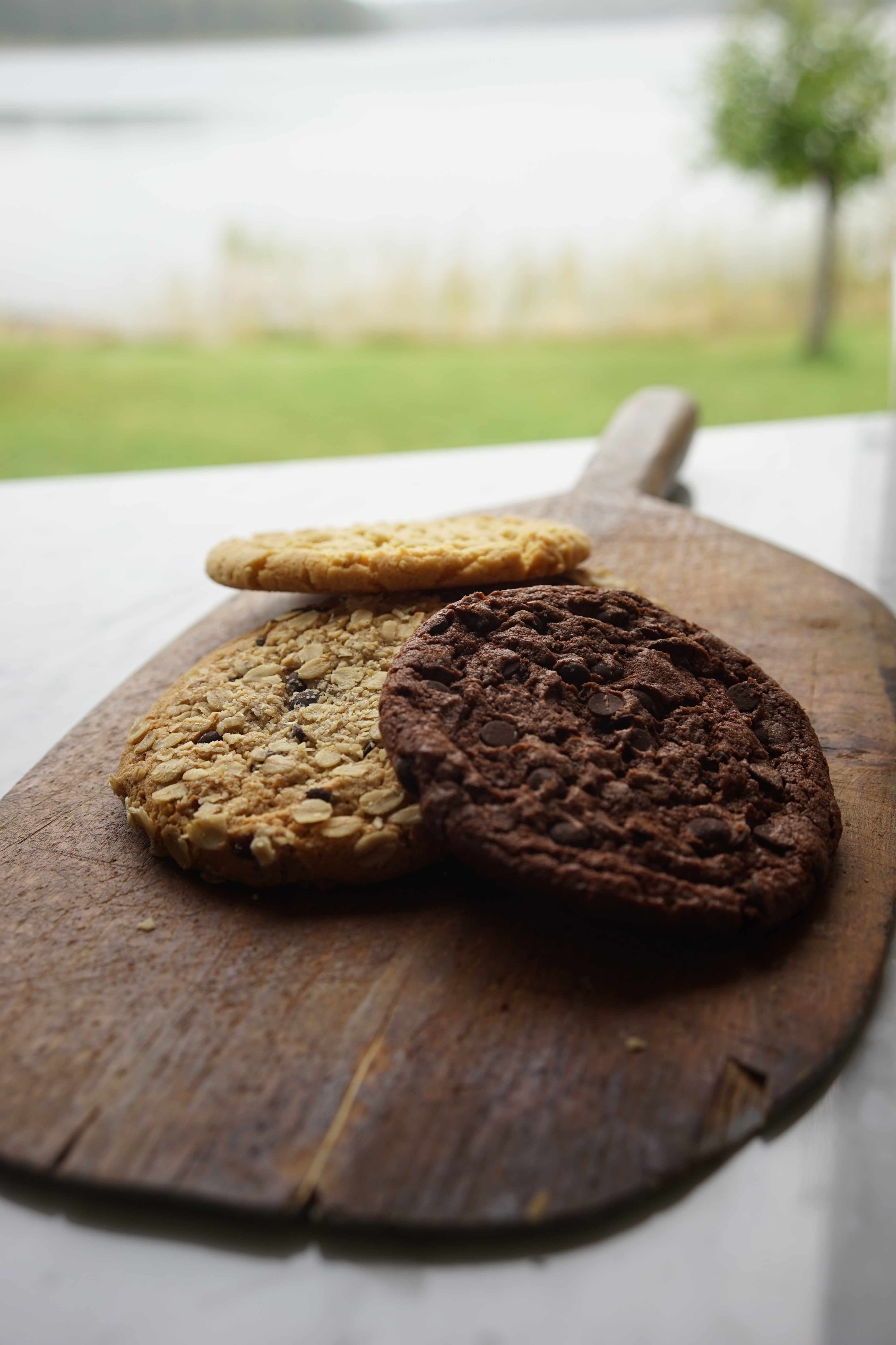 Foto: Espresso House Tre cookies ligger på en skärbräda i trä på ett vitt bord. Två ljusbruna och en mörkbrun.