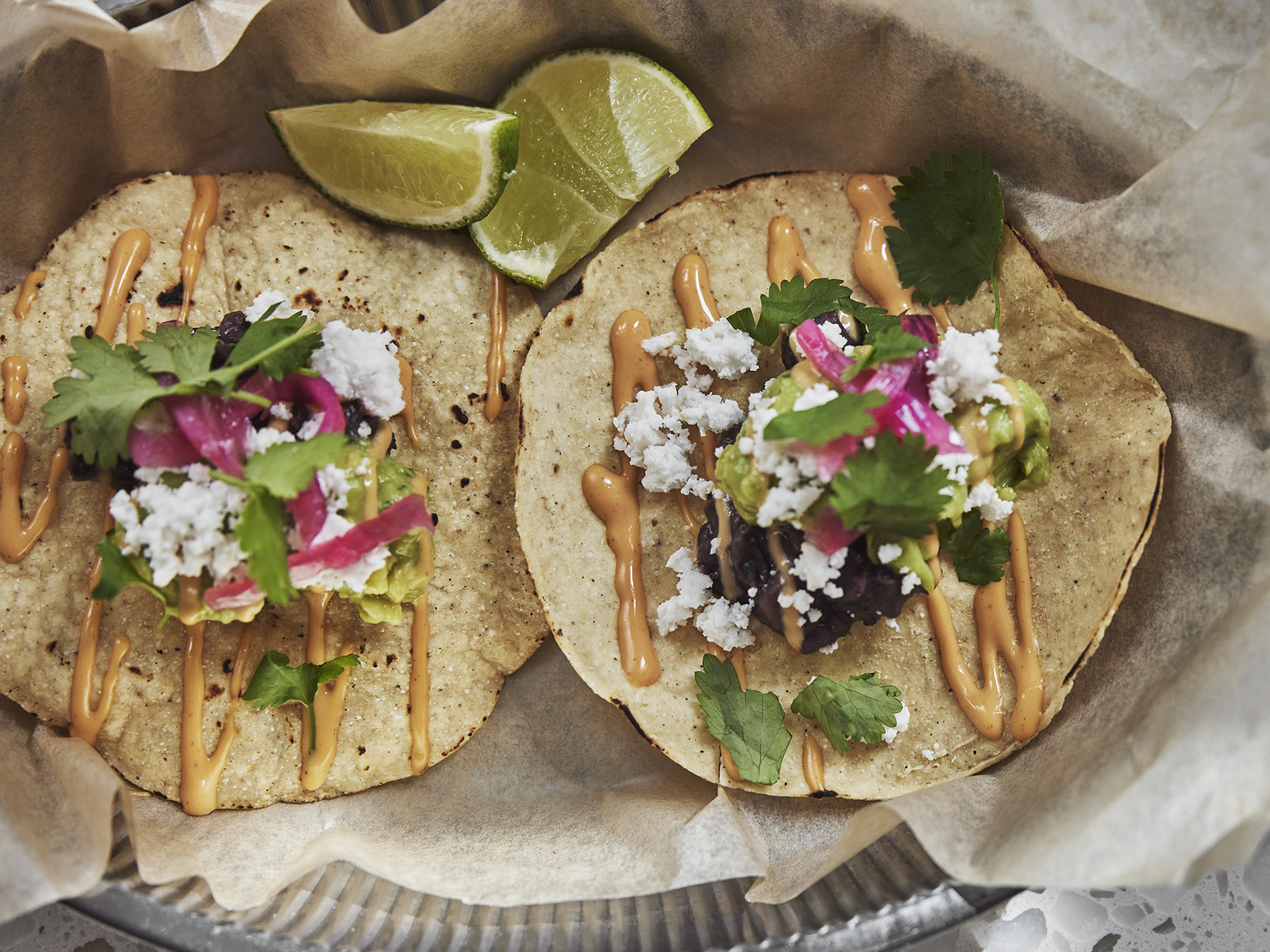 Foto: Hannes Söderlund/El Taco Truck Två, små tacobröd fyllda med bl.a. rödlök, avocado, koriander, vegansk ost och sås, liggandes på bakplåtspapper bredvid limeklyftor.