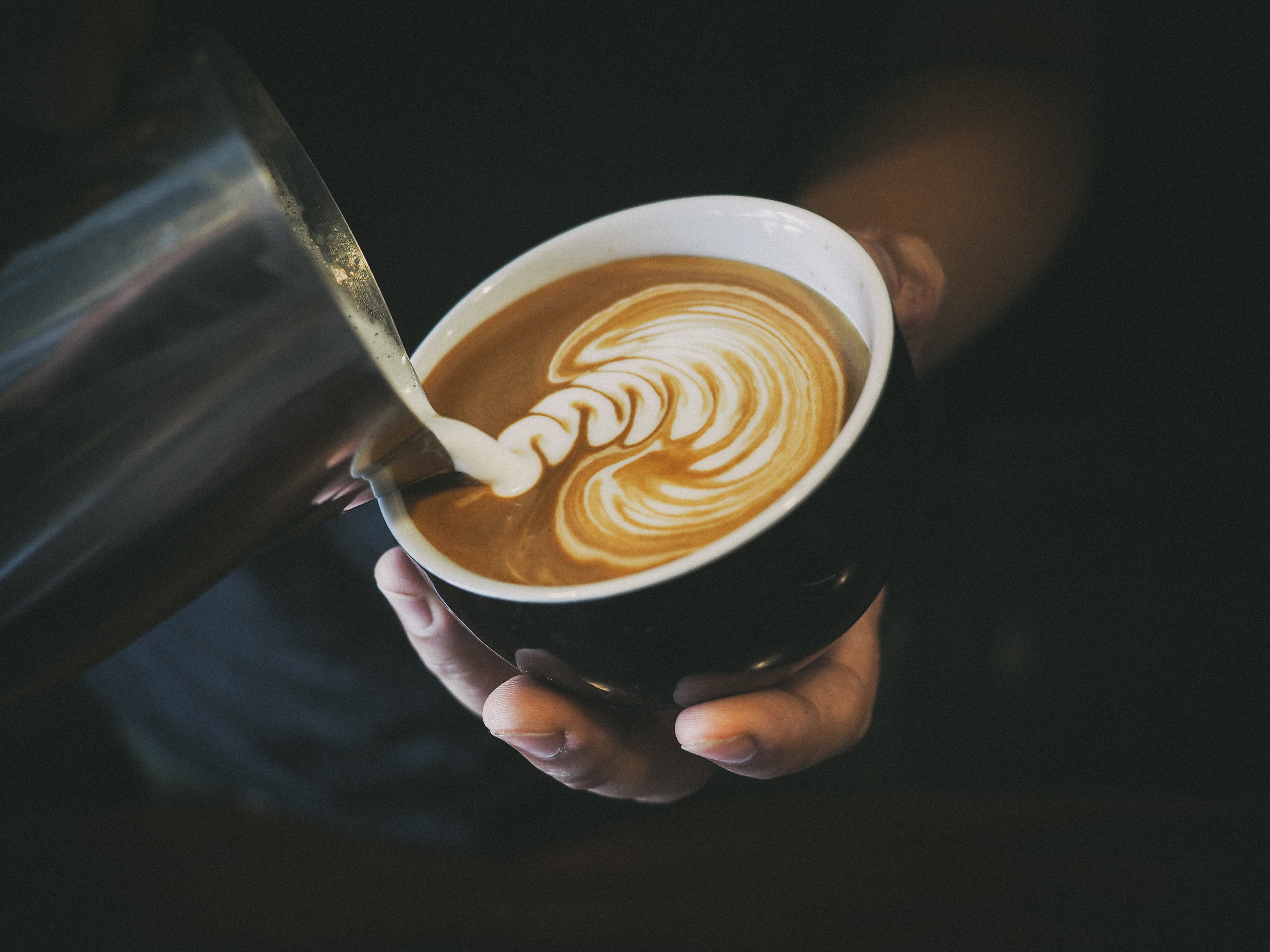 Foto: StudioByTheSea/Shutterstock "Mjölk" hälls i kopp med kaffe. Koppen hålls i en hand i dovt belyst miljö.