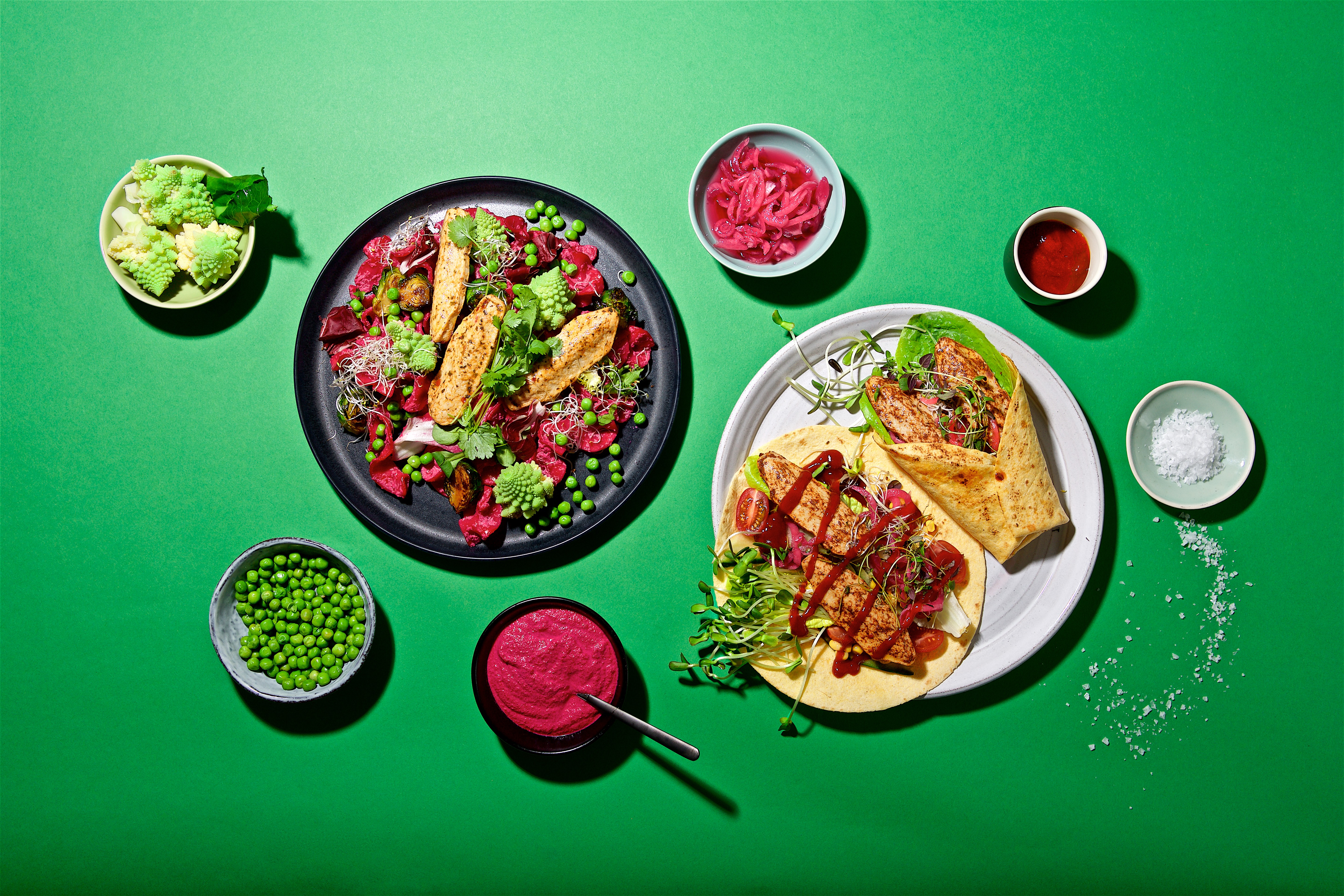 Produktfoto: Quorn veganska filléer i en sallad på en tallrik och en i wrap på en annan tallrik uppdukat på ett grönt bord med tillbehör.