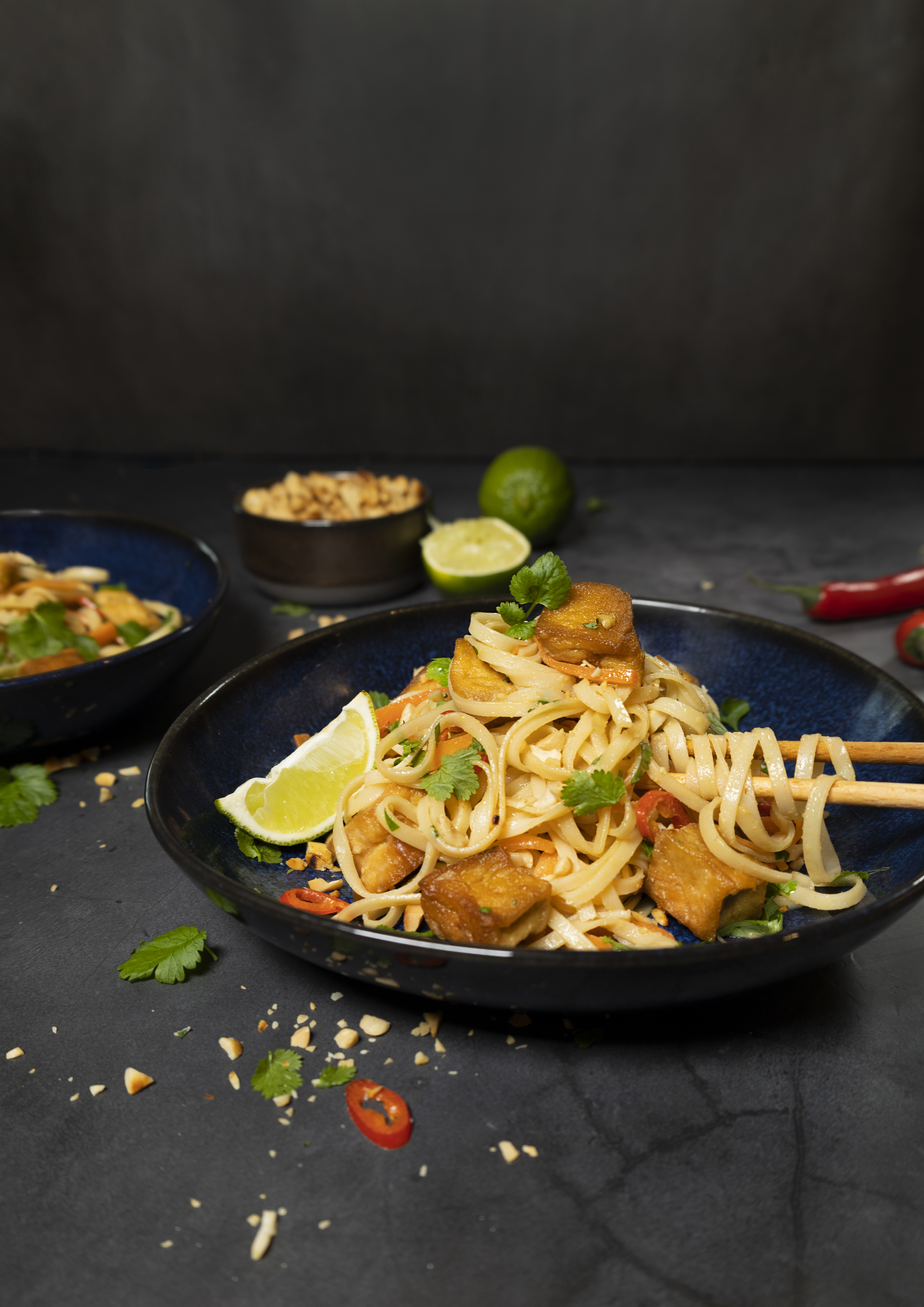 Produktfoto: Yipin Veganska pad thai med tofu i en mörkblå djuptallrik mot en mörkgrå bakgrund.