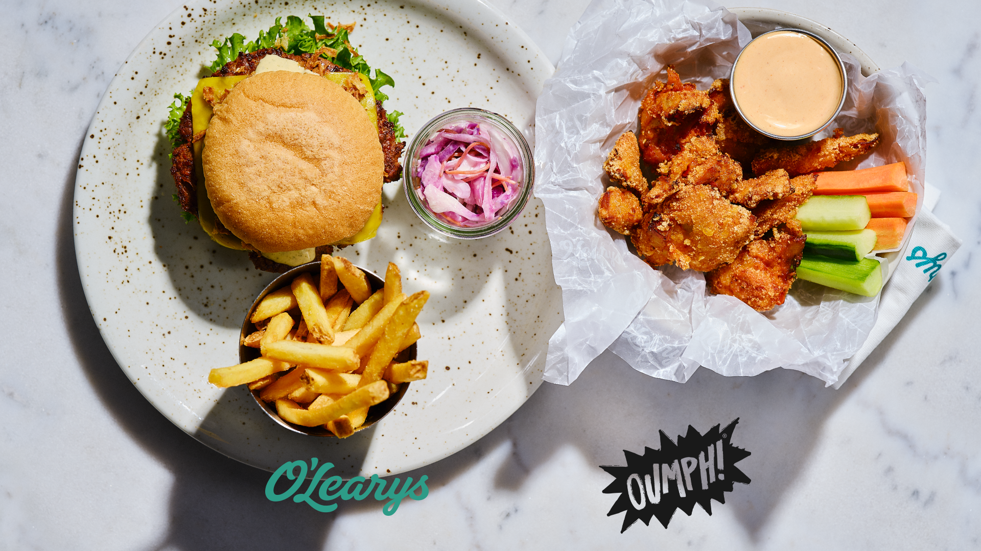 Produktfoto: O'Learys, Oumph! En tallrik med hamburgare, pommes och coleslaw och en med veganska chicken wings, grönsaker och dip. Bild tagen uppifrån.