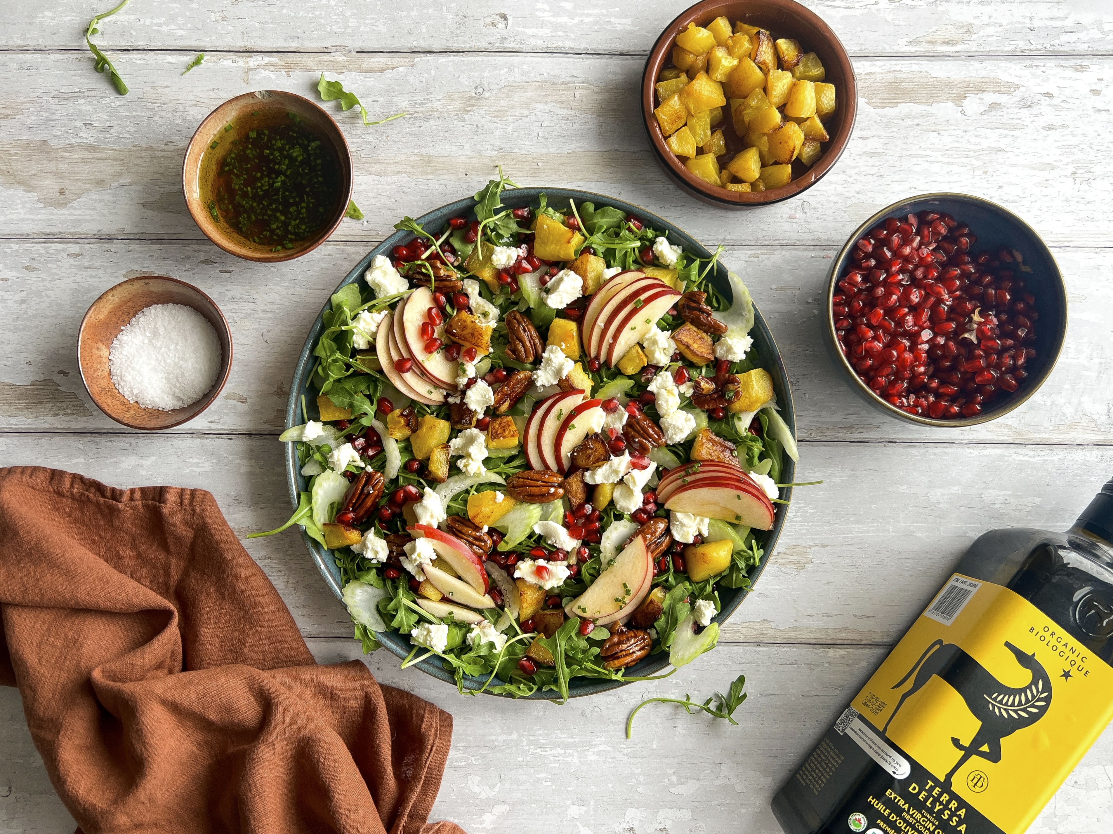 Produktfoto: Terra Delyssa Sallad i en skål på ett vitt bord med granatäpplekärnor, mango, olivolja och en brun kökshandduk runt om tallriken.