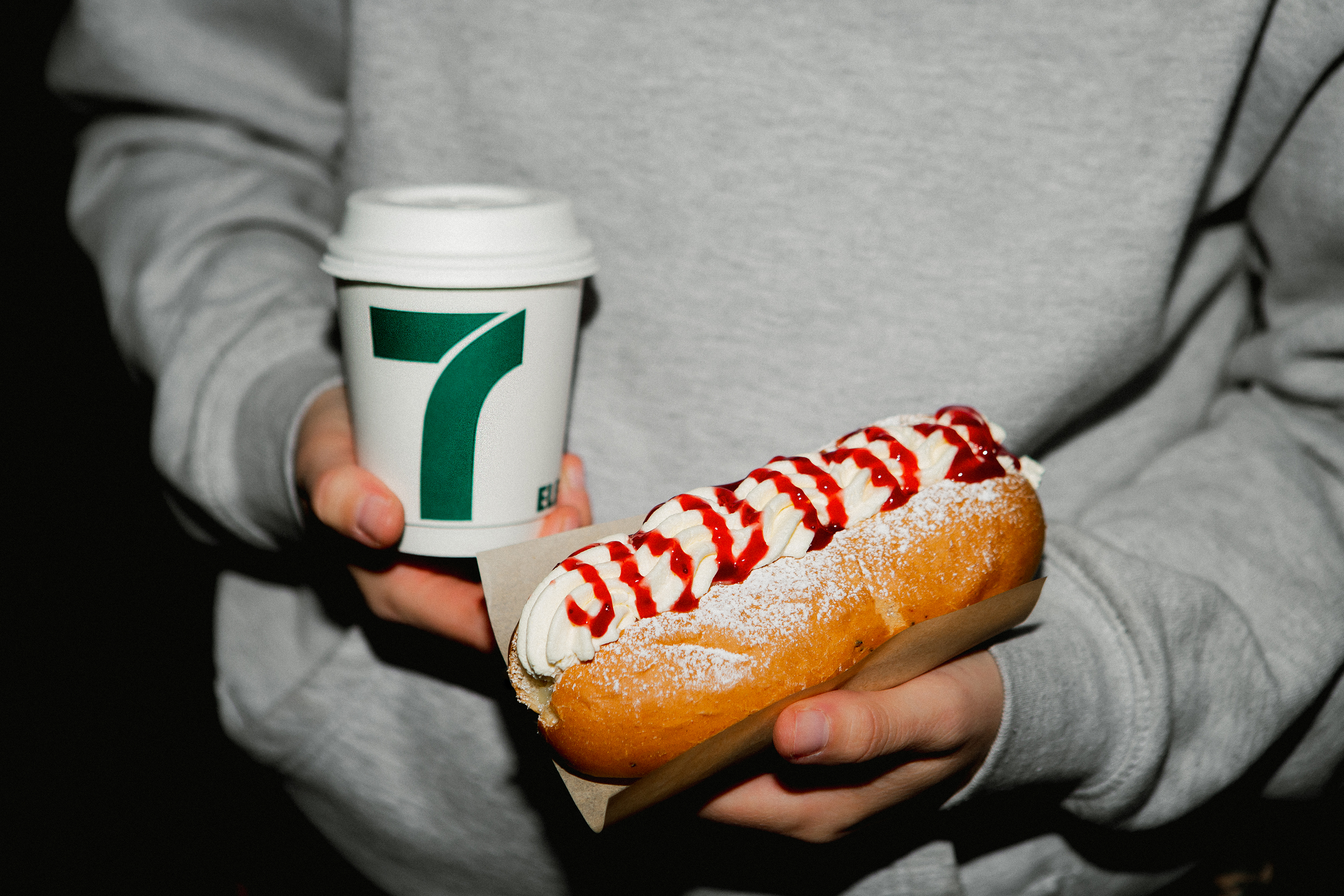 Produktfoto: 7-Eleven Man som håller en pappmugg med kaffe i andra handen och en semmelbakelse i korvbröd i den andra