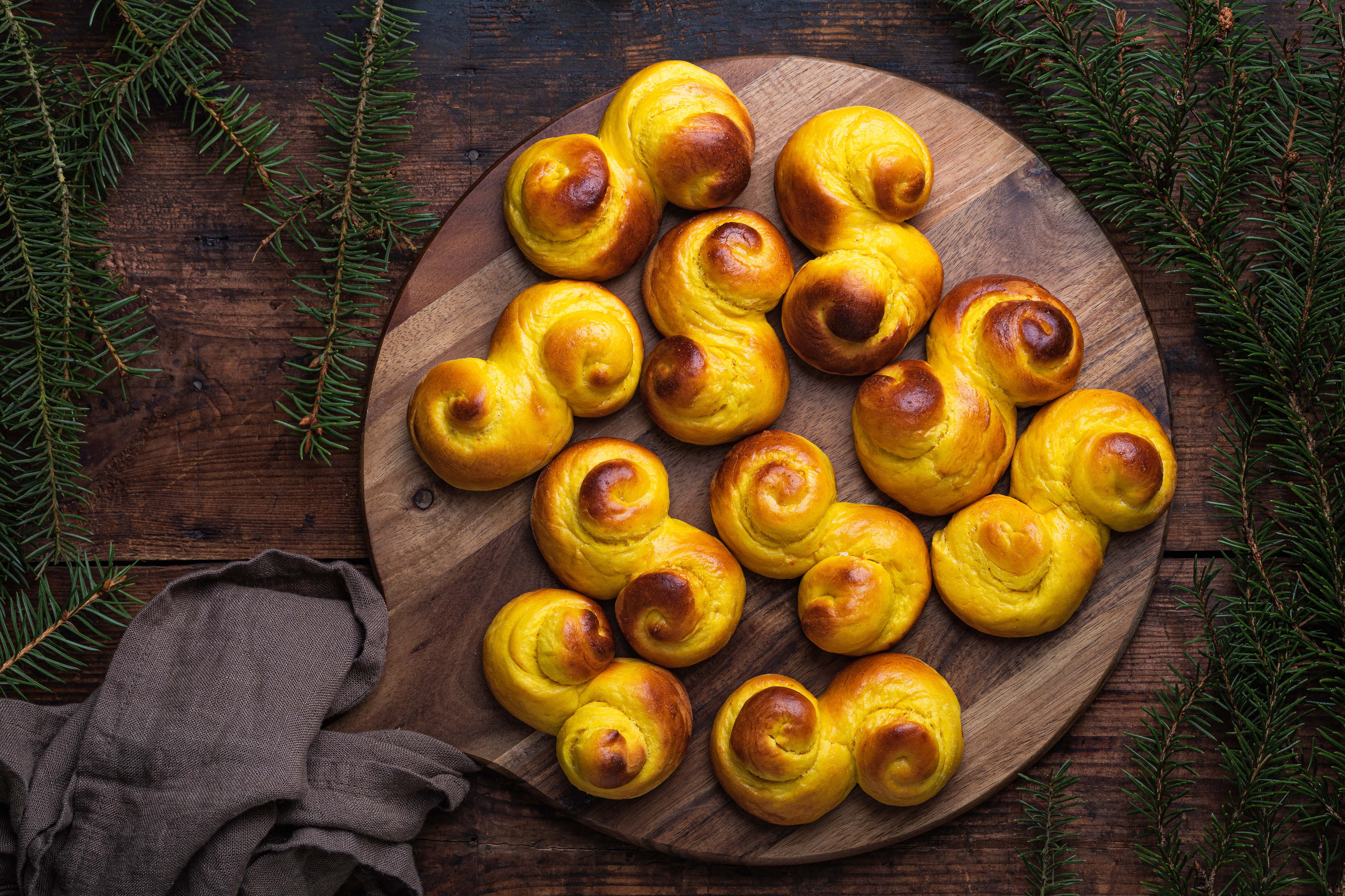 Foto: Angela Kotsell/Shutterstock Freshly baked homemade Swedish traditional s-shaped saffron buns, also known as lussebullar or saffransbröd. Seen from above flat lay on a dark wooden table surrounded by spruce branches.