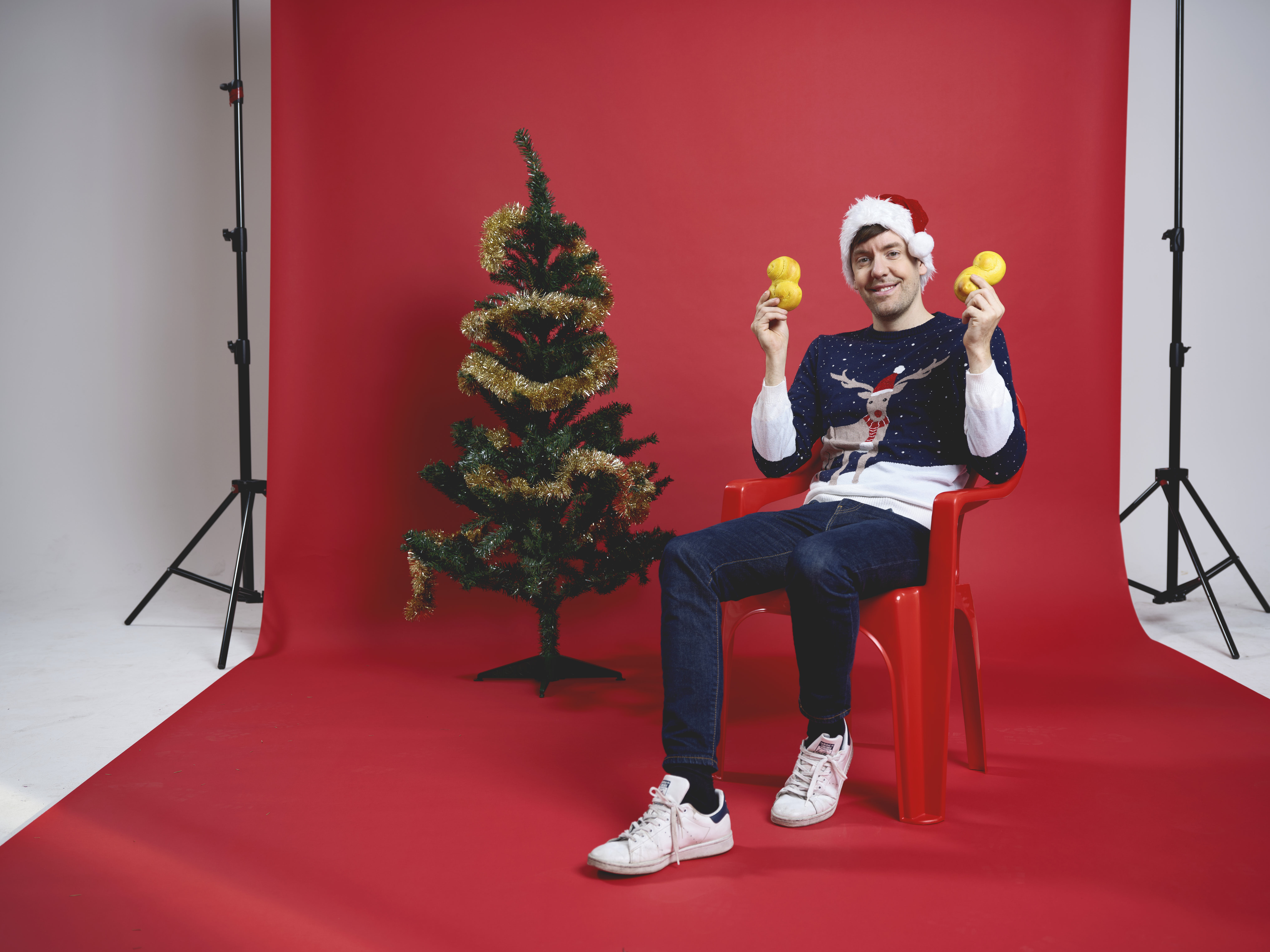 Foto: Oskar Omne En man som sitter på en stol framför en röd fotovägg. Han håller upp lussebullar, har en jultröja på sig och sitter bredvid en julgran.
