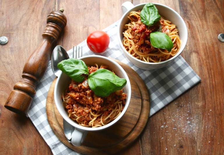 Två skålar med pasta bolognese på en rutig handduk, bredvid ligger en tomat och en pepparkvarn