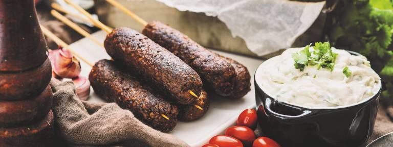 kebabspett på en vit skärbräda, bredvid spetten står en skål med tzatziki  