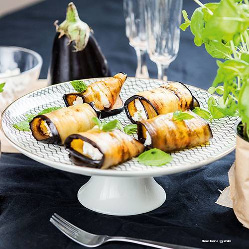Auburginerullar med vegansk fetaost på ett vitt tårtfat på ett mörkt bord. 