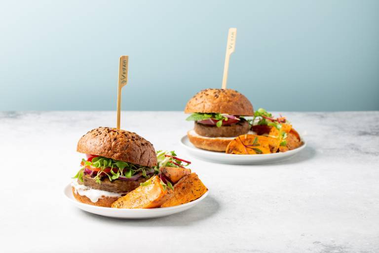 Köttfri växtbaserad hamburgare serveras med sötpotatisklyftor, grön mixsallad och vit sås på grått bord nära blå bakgrund. 