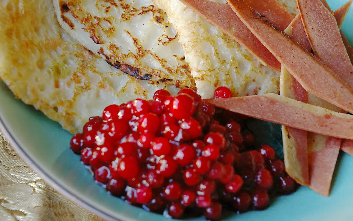 Raggmunkar med stekt bacon och rårörda lingon på ett blått fat.