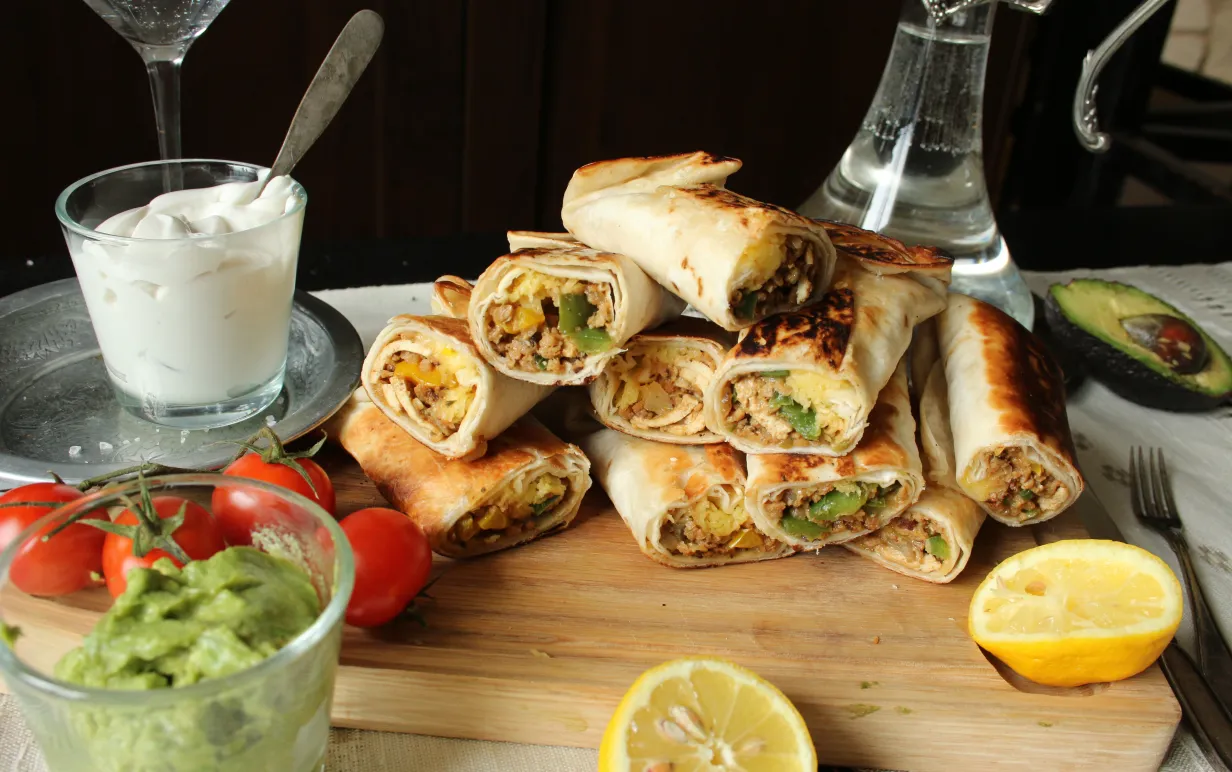 Chimichangas i en hög på en skärbräda, bredvid står en skål med vegansk créme fraiche och en skål med guacamole