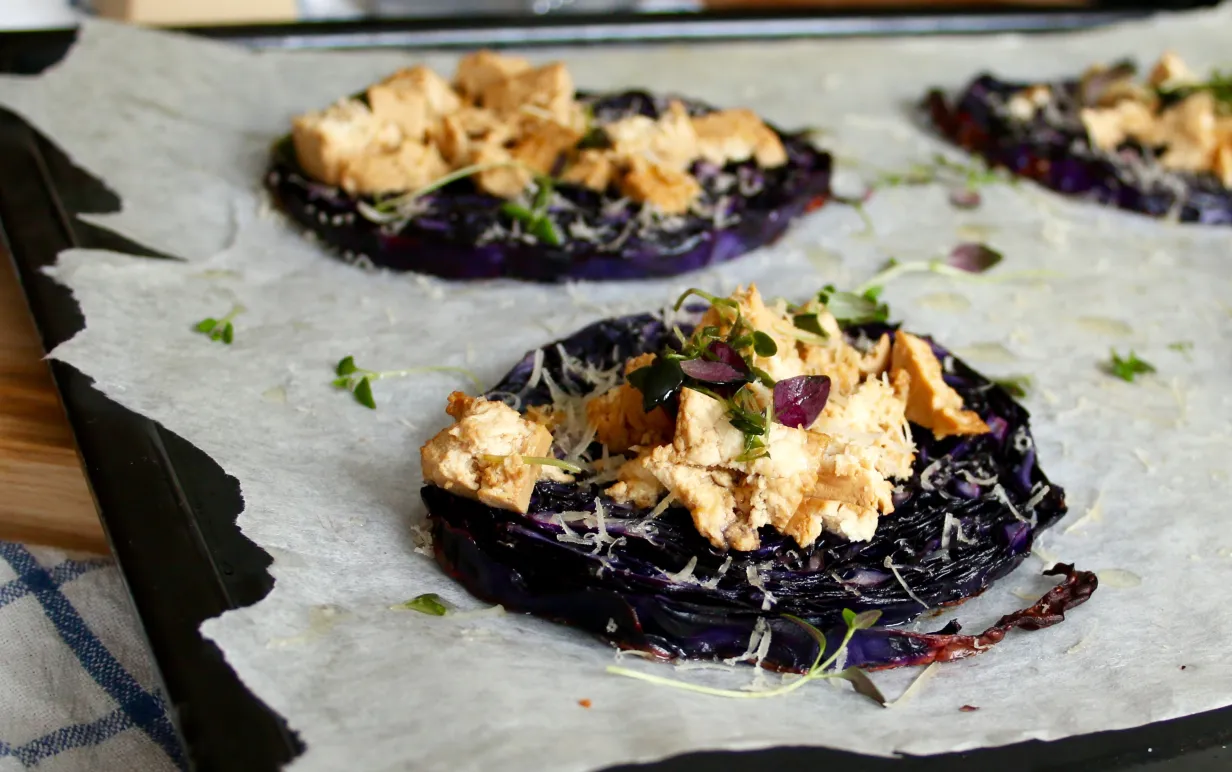 Rödkålskivor toppade med tofu och parmesan på ett bakplåtspapper.