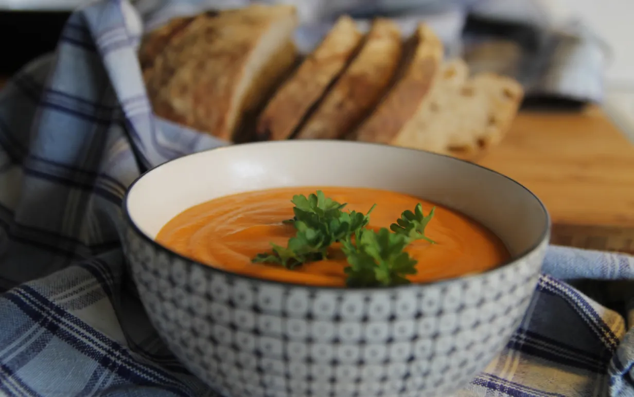 Sötpotatissoppa i en skål på en rutig handduk på ett bord. Det ligger bröd bredvid handduken.