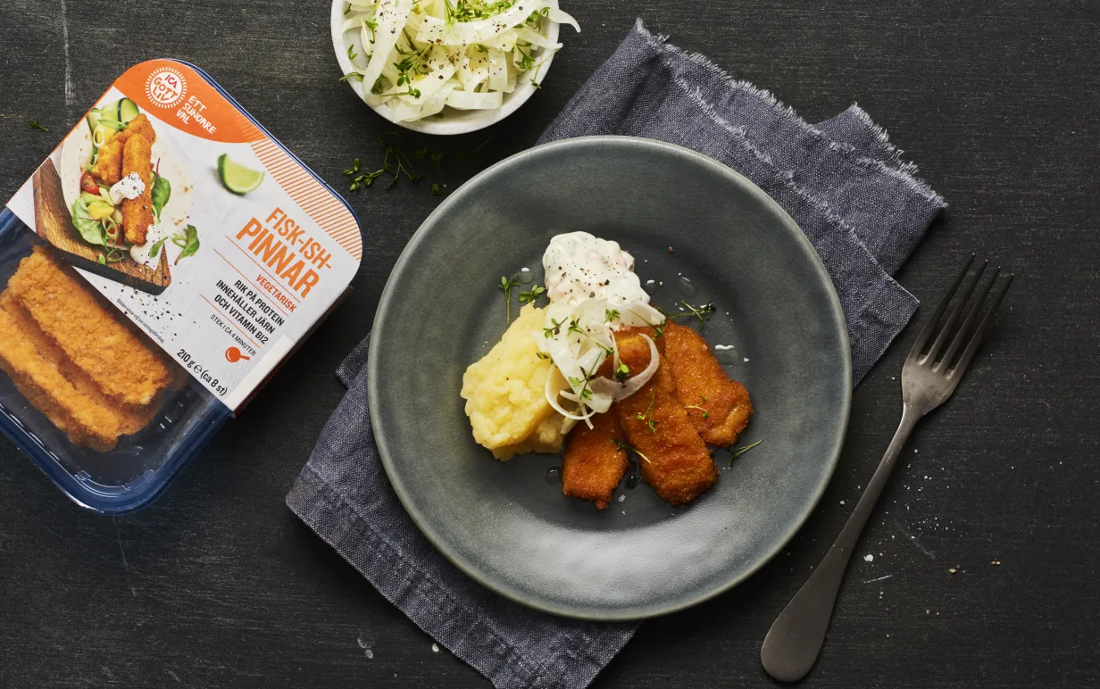 Svart tallrik med veganska fiskpinnar, potatismos och sås bredvid en förpackning veganska fiskpinnar.