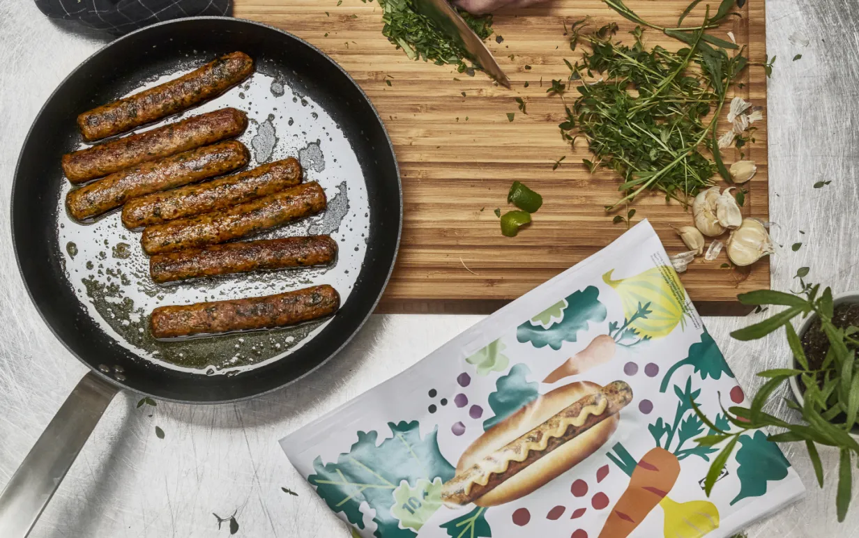 Händer hackar gräslök och det ligger ett par veganska korvar på en panna intill