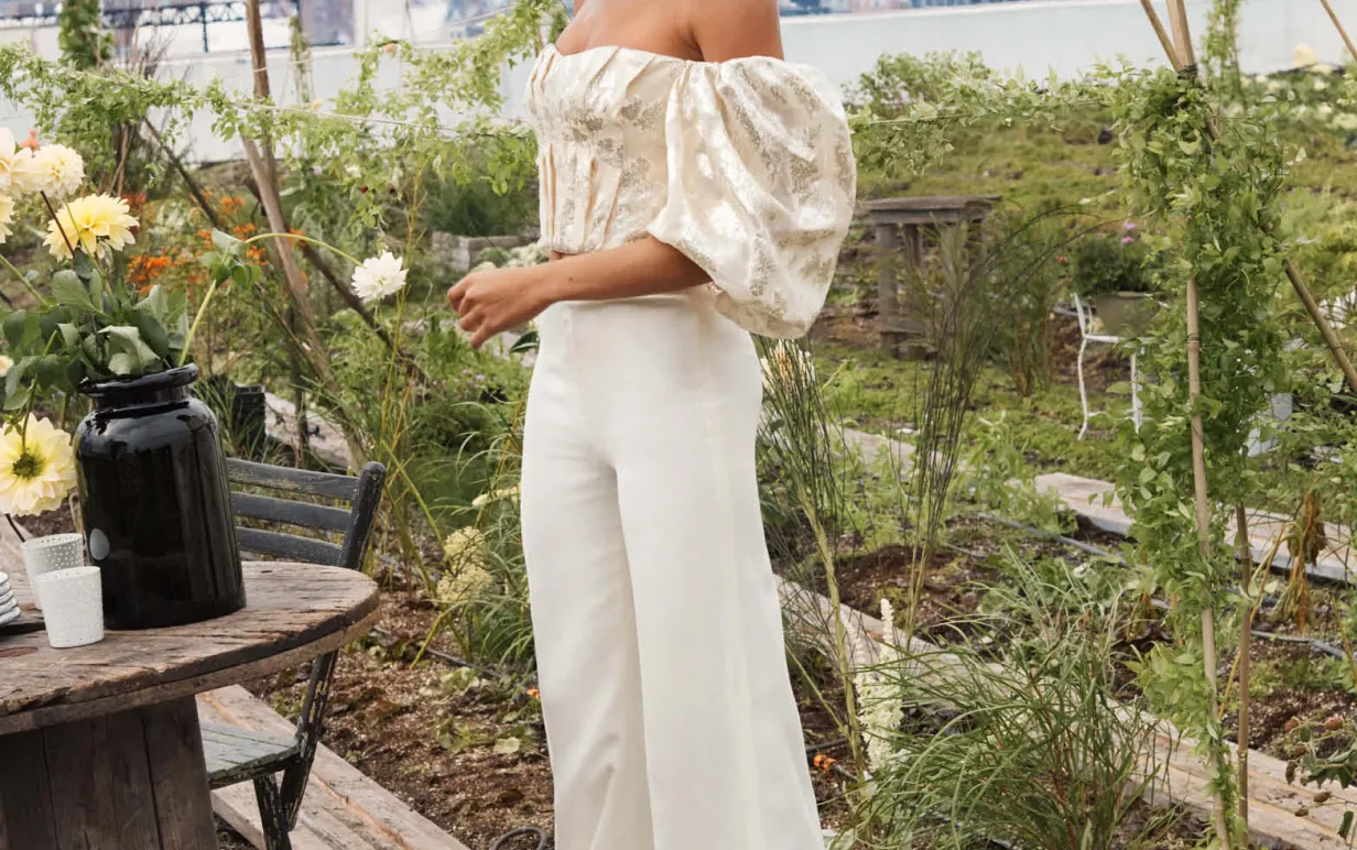 Person med vita byxor och vit topp står i en trädgård.