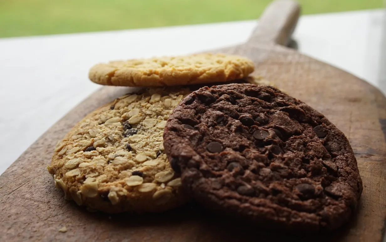 Tre cookies ligger på en skärbräda i trä på ett vitt bord. Två ljusbruna och en mörkbrun.