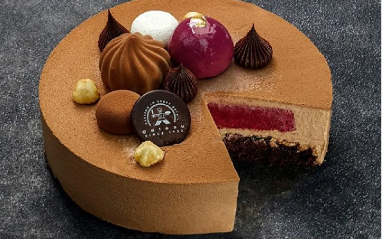 En brun rund tårta där en bit har tagits så att innehållet med svartvinbär och mousse syns. Toppat med dekorationer från Gateau