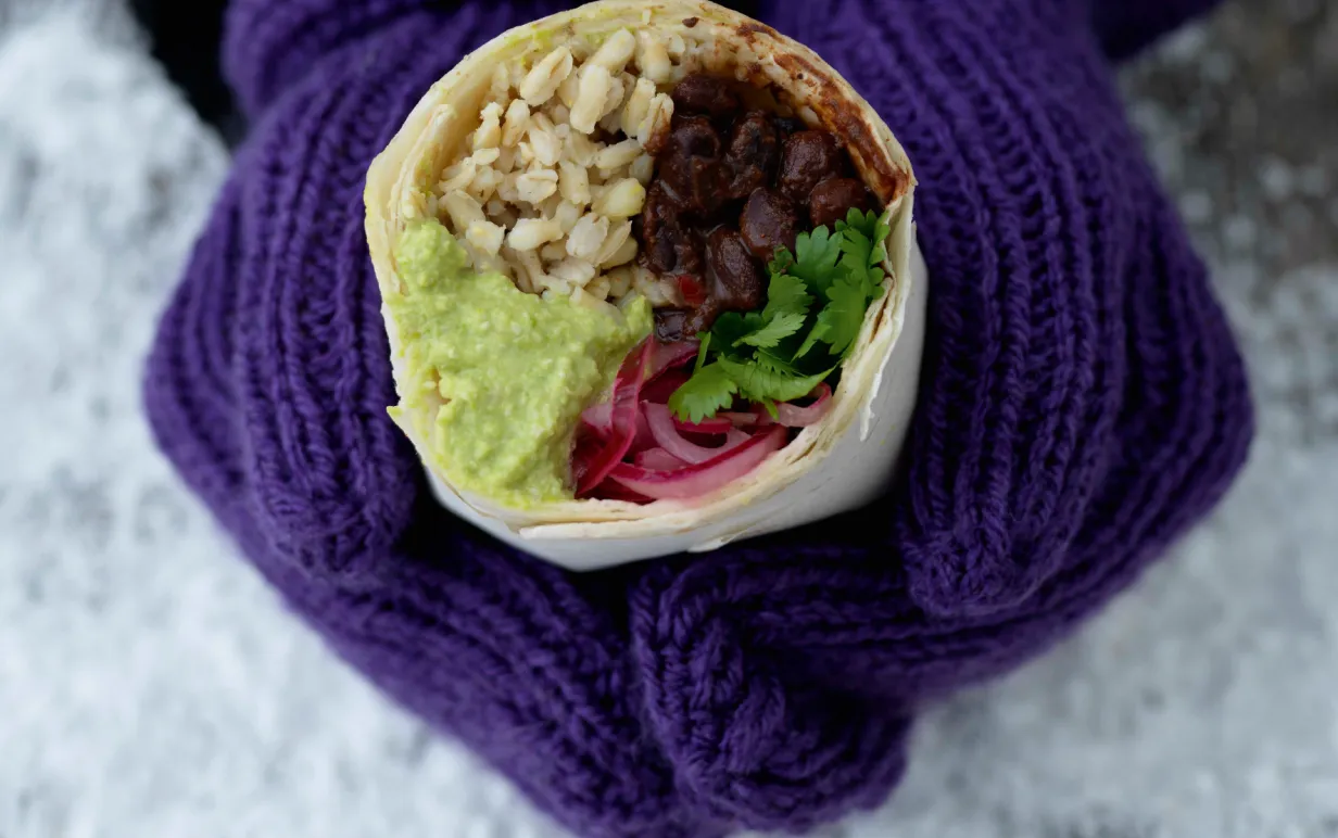 händer med lila vantar håller i en burrrito med krämiga svarta bönor, guacamole och ris