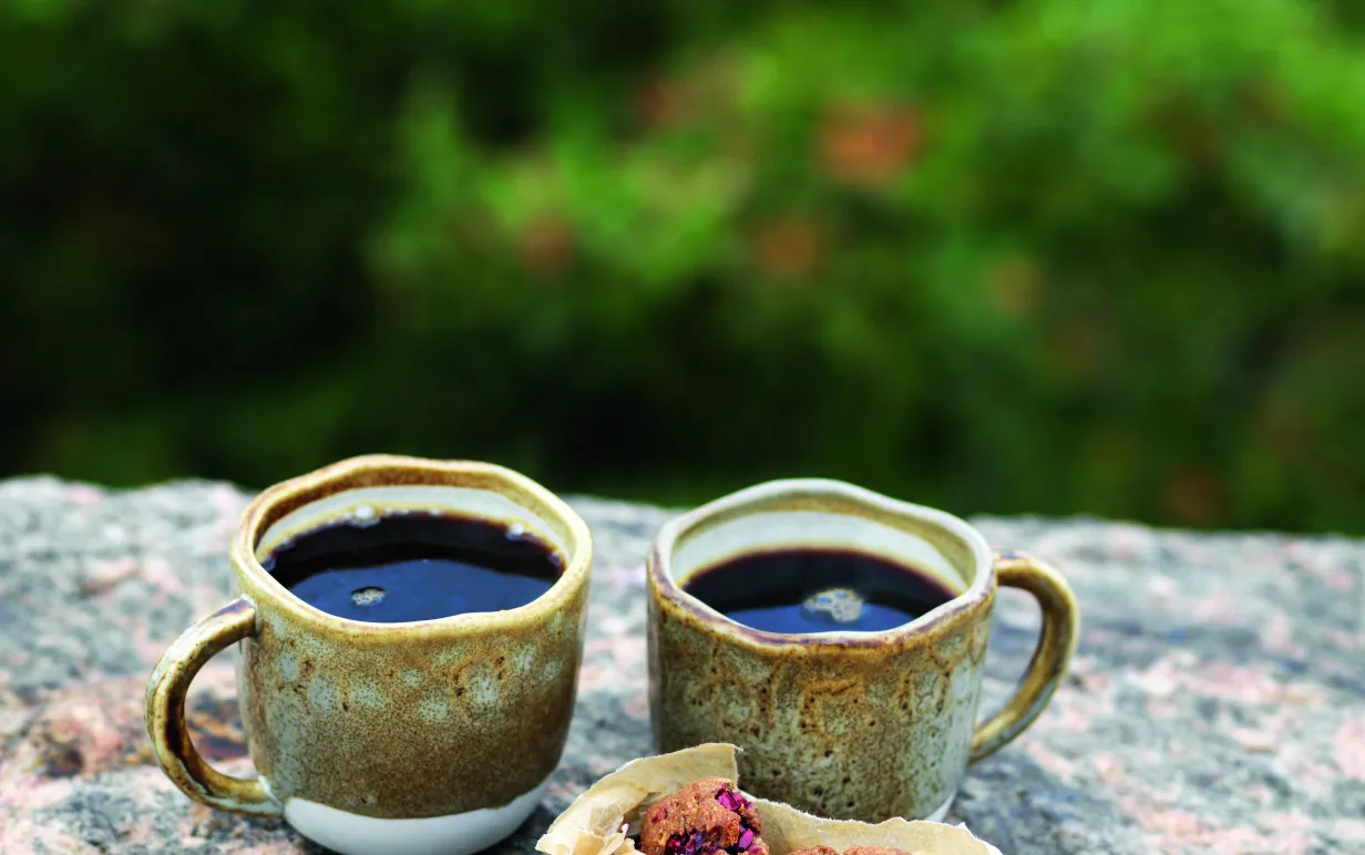två tahinicookies på en sten med två kaffekoppar bakom