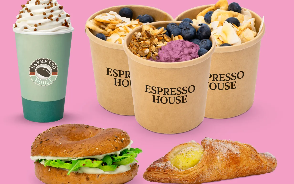 Cafe Latte i pappersmugg, tre små papperskoppar med overnight oats resp. chiapudding med färgglada toppings, avokadobagel och croissant mot rosa bakgrund.