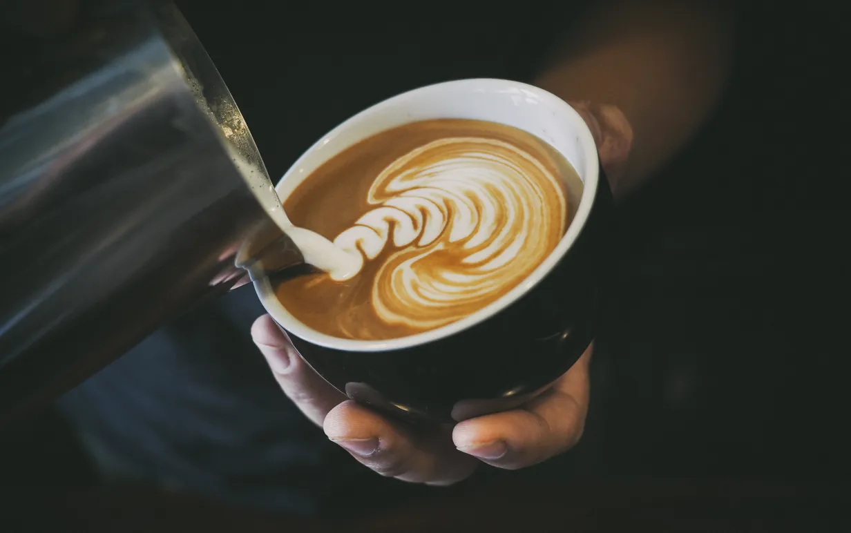 "Mjölk" hälls i kopp med kaffe. Koppen hålls i en hand i dovt belyst miljö.