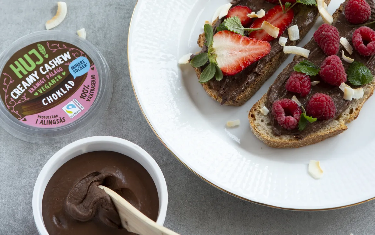 HUJ Creamy Cashew Choklad i öppnad förpackning till vänster. Till höger på macka med bär och kokos.