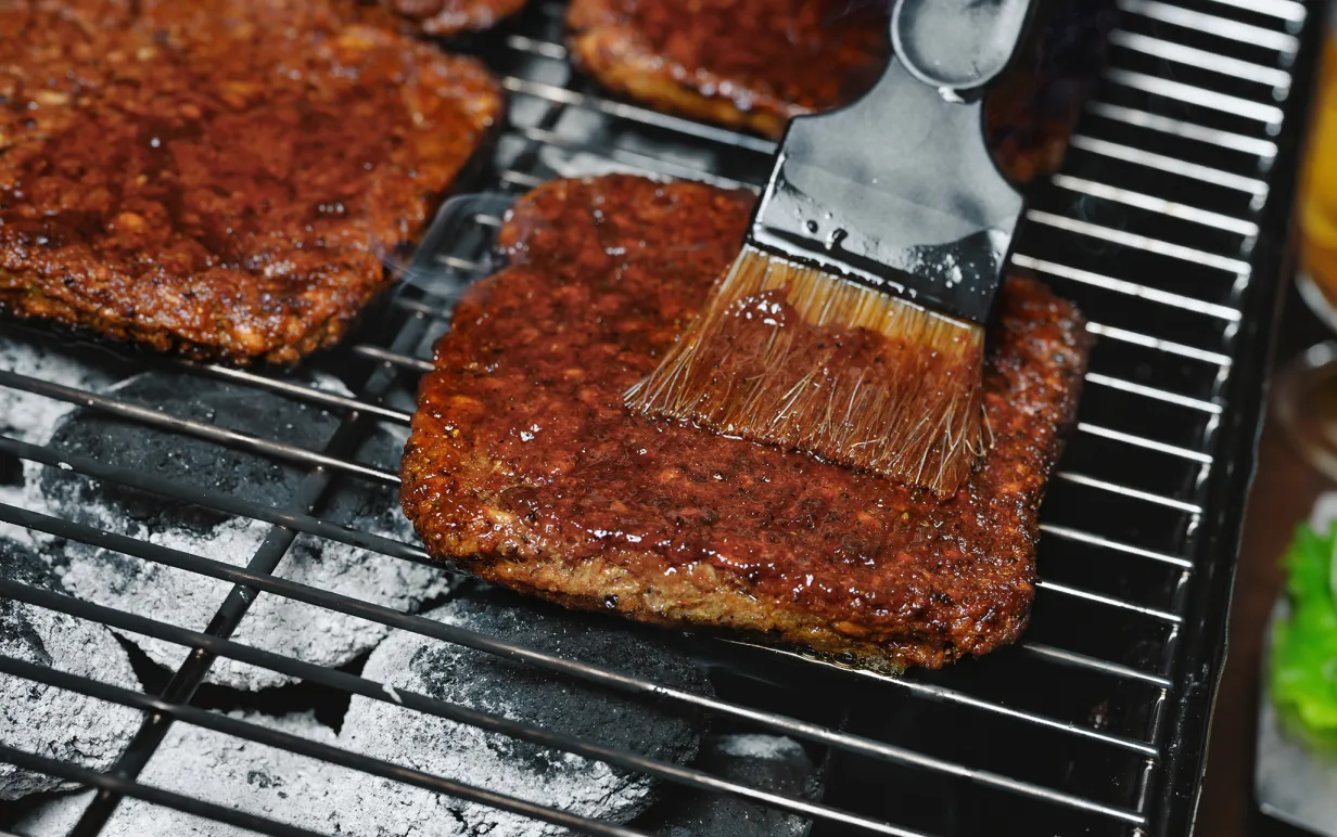 Fyrkantiga veganska burgare på en grill som blir penslad med grillolja.
