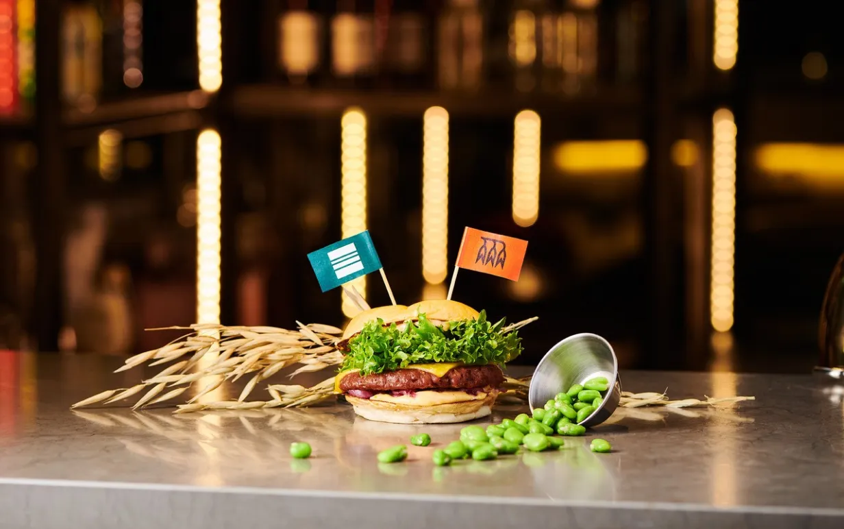 En växtbaserat burgare på ett bord med två flaggor i brödet dekorerat med havre och sojabönor runt burgaren.