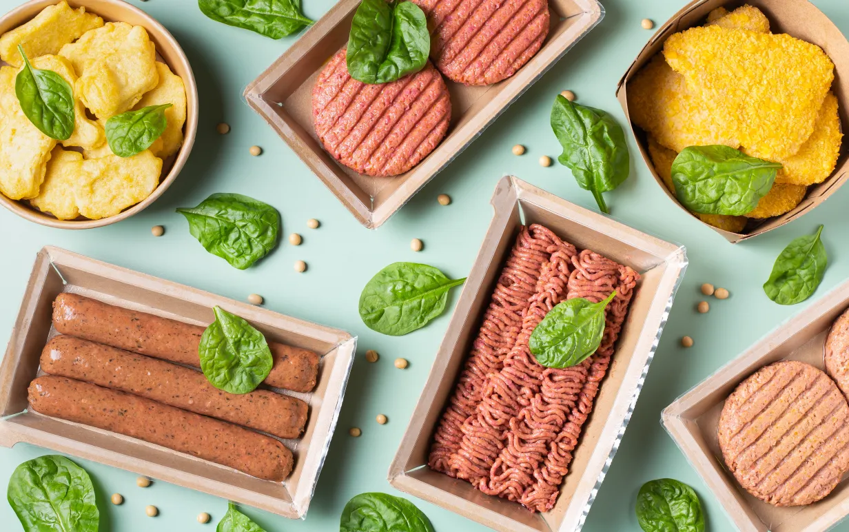 Flera olika växtbaserade köttsubstitut upplagda på ett mintgrönt bord med basilikablad utspridda.