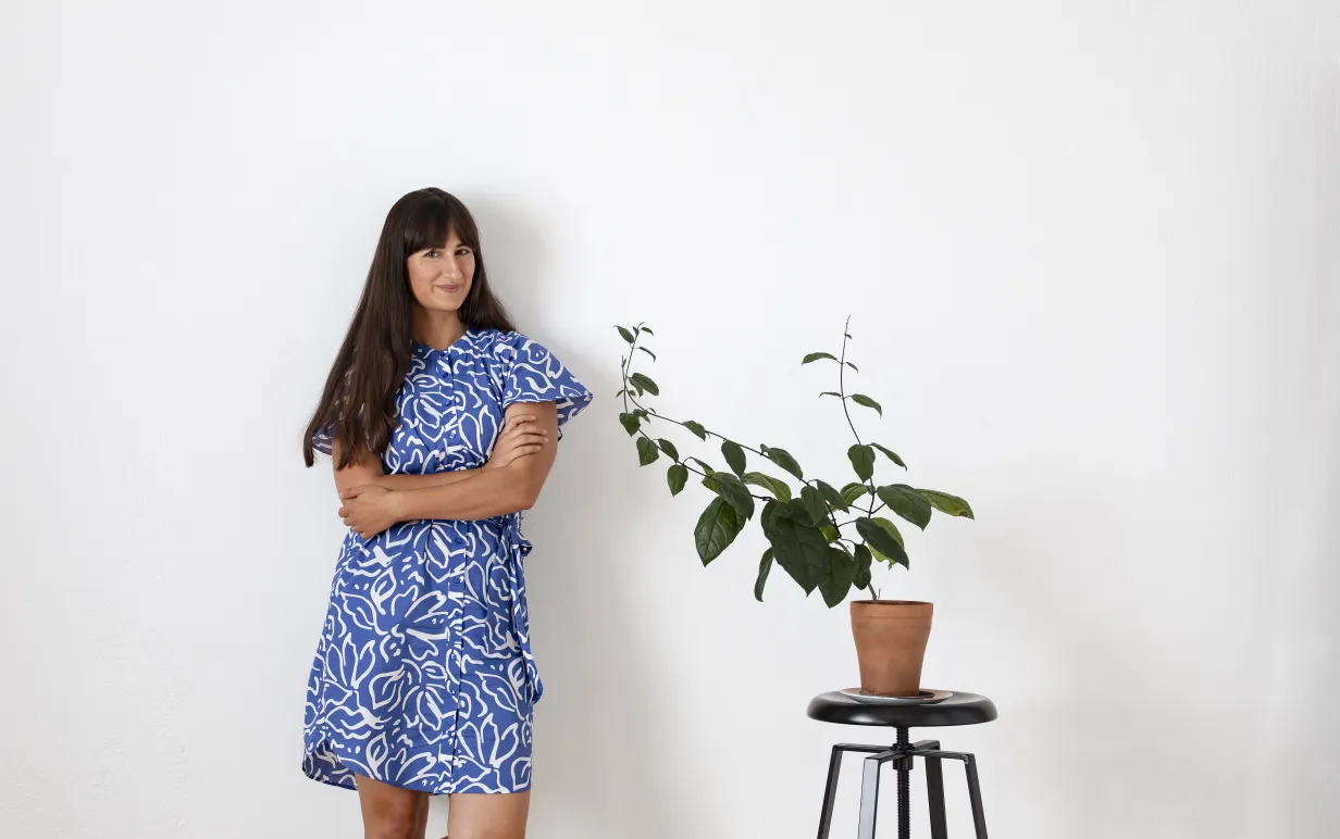 Mörkhårig kvinna i blå klänning bredvid en grön krukväxt mot en vit vägg.