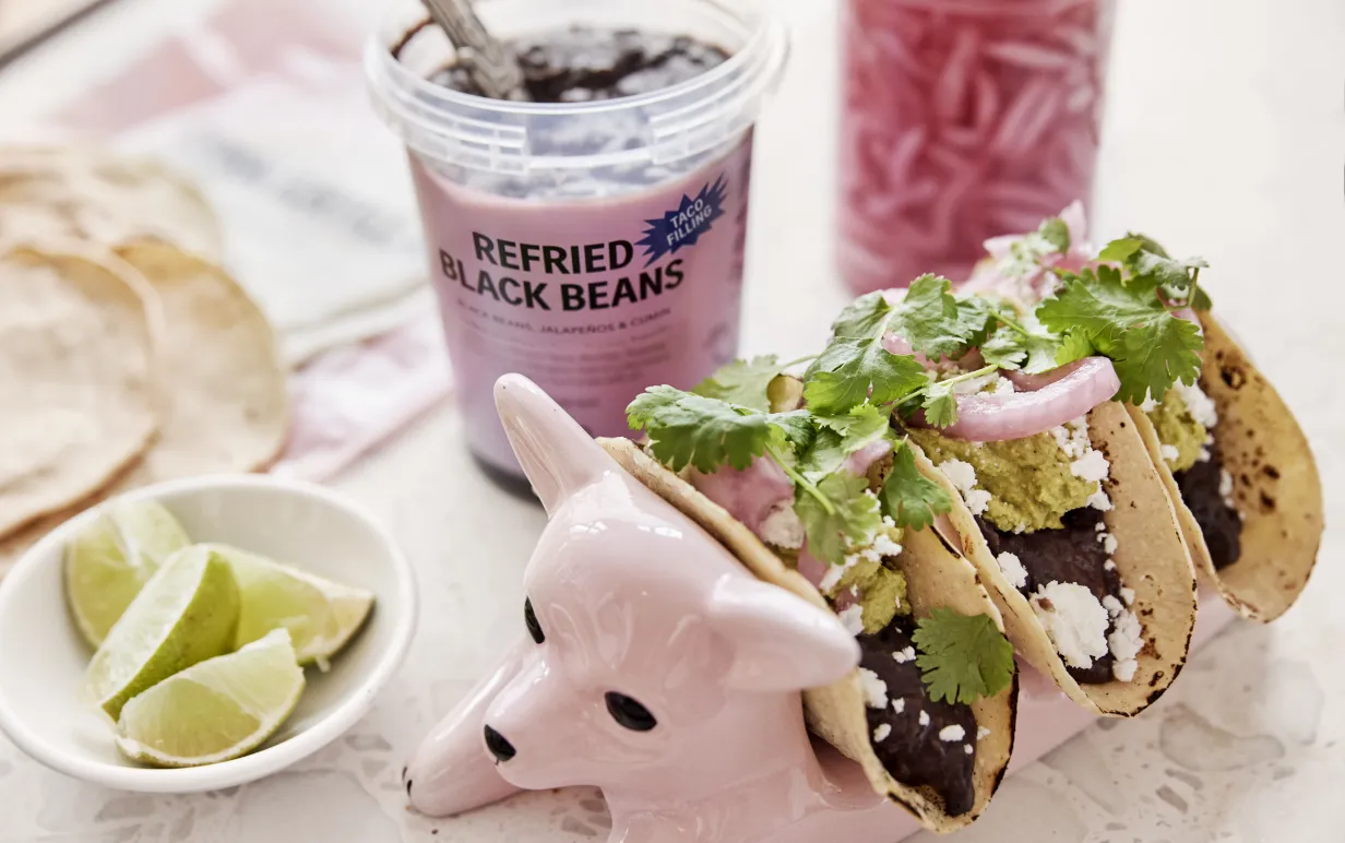 Mjuka fyllda veganska tacos uppställda i en keramikhund med läsk, lime och picklad rödlök på sidan om.