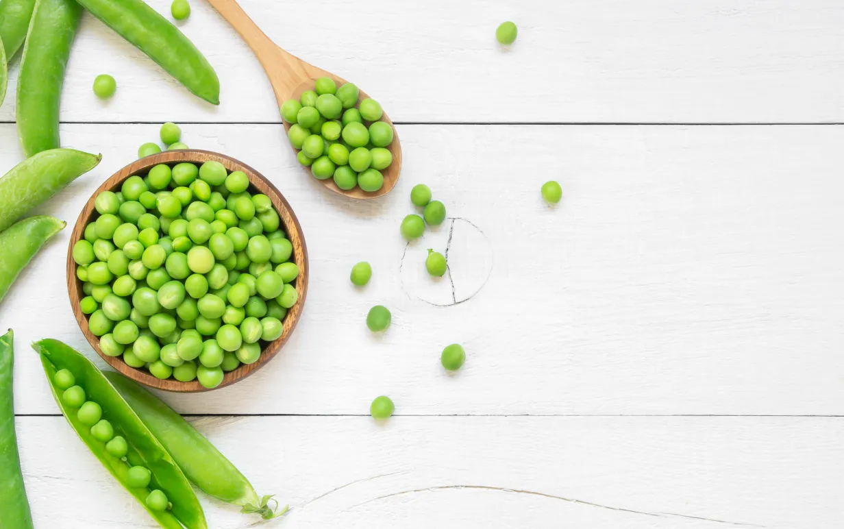 gröna ärtor i en träskål bredvid en träsked och ärtskidor på ett vitt bord.