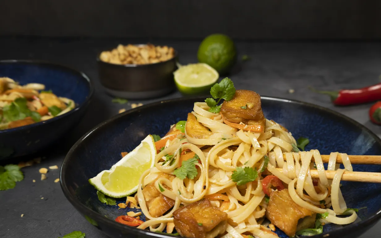 Veganska pad thai med tofu i en mörkblå djuptallrik mot en mörkgrå bakgrund.