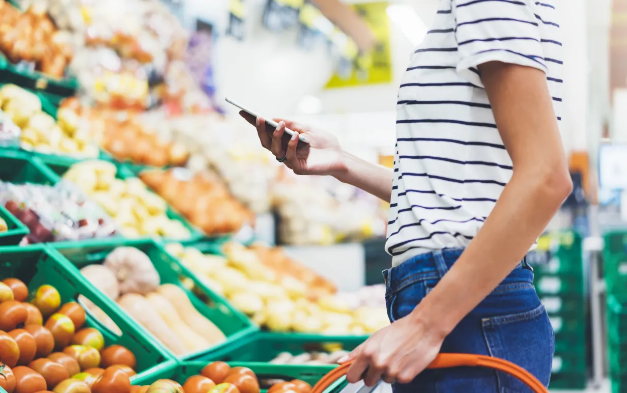 Ung kvinna handlar hälsosam mat i stormarknad suddig bakgrund. Kvinnliga händer köper naturprodukter med smart telefon i butik. Hipster på livsmedelsbutiker med hjälp av mobil. Person som jämför priset på produkter