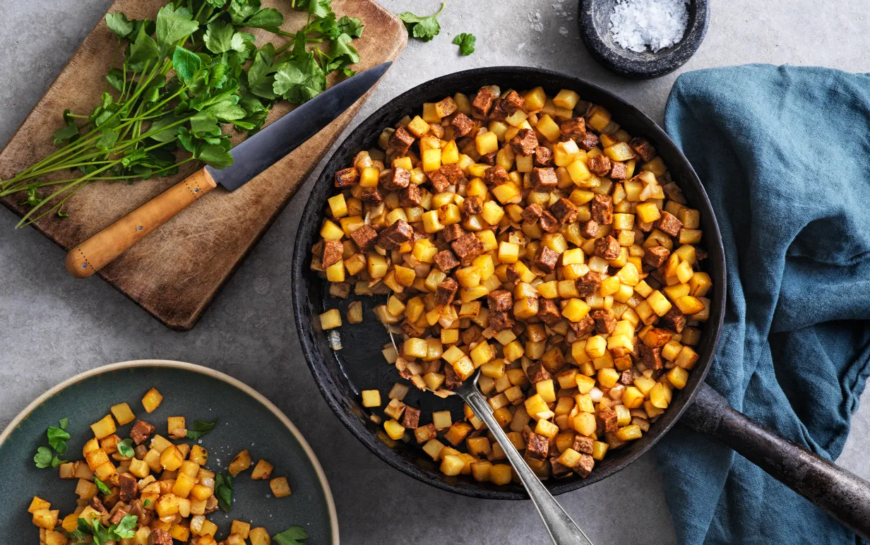 Vegansk pytt i en stekpanna bredvid en tallrik med pytt och en skärbräda med örter