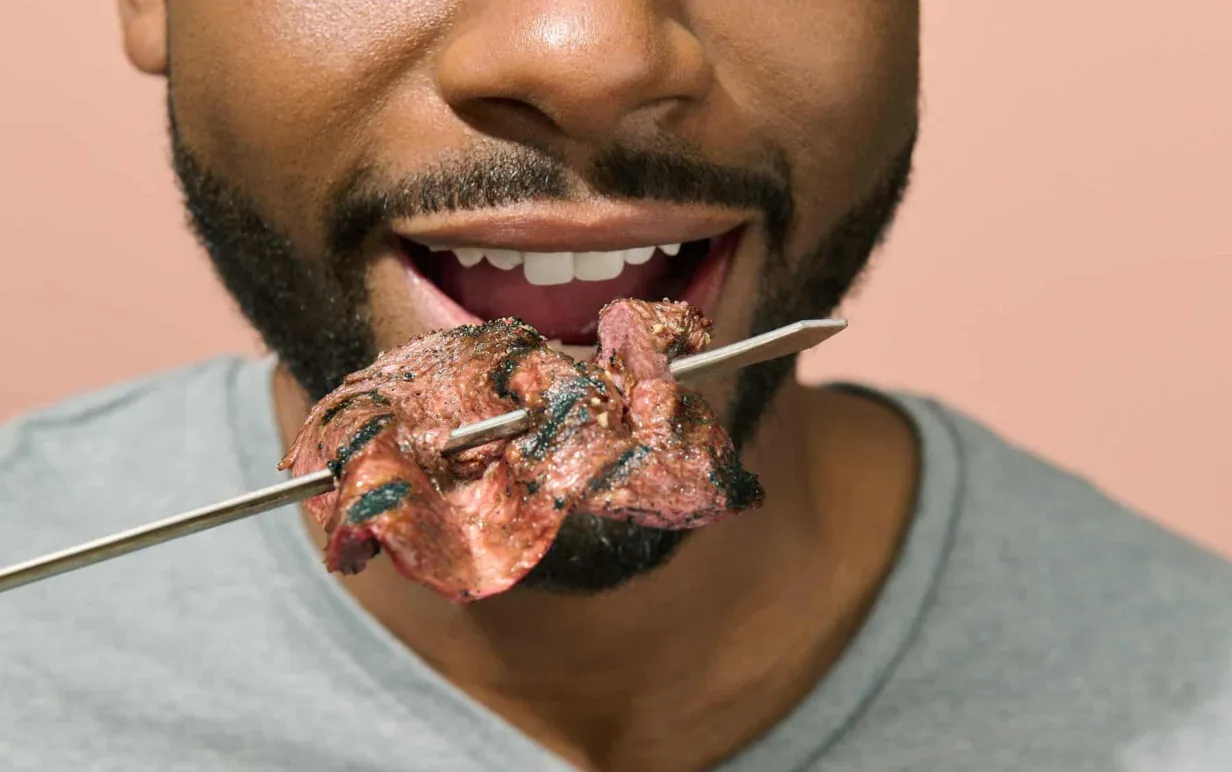 Bild på en mans mun med ett grillspett med kött framför sig