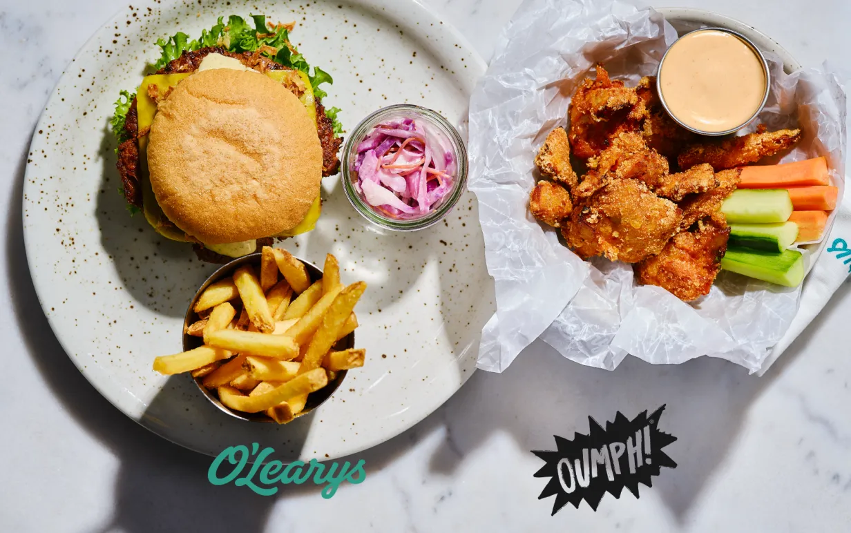 En tallrik med hamburgare, pommes och coleslaw och en med veganska chicken wings, grönsaker och dip. Bild tagen uppifrån.