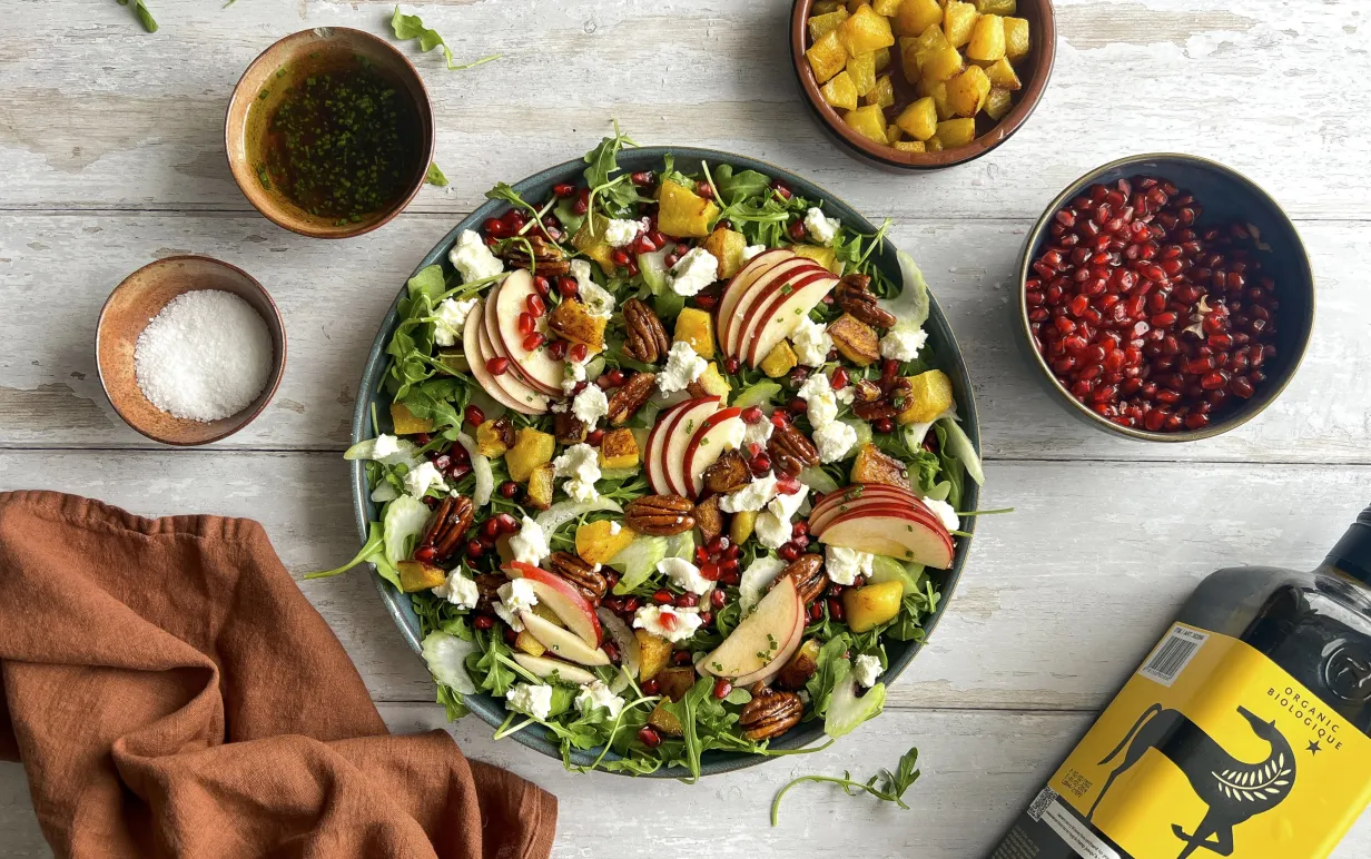 Sallad i en skål på ett vitt bord med granatäpplekärnor, mango, olivolja och en brun kökshandduk runt om tallriken.