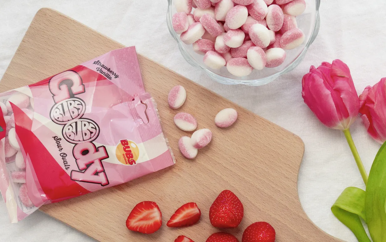 En påse godis öppnat och utspridd på en skärbräda i trä med jordgubbar och röda tulpaner runt