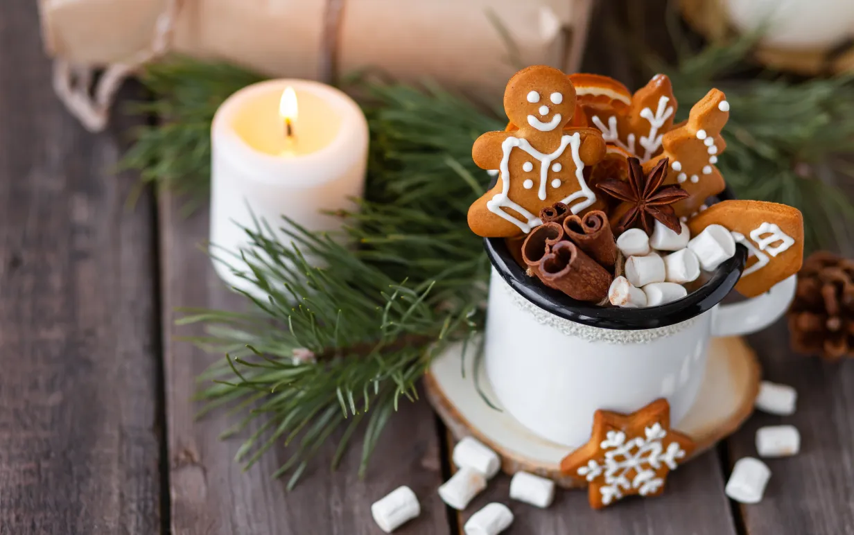 Hot winter drink in a white mug: cozy home composition with homemade gingerbread cookies, tree branch. Wooden background, christmas lights and candles