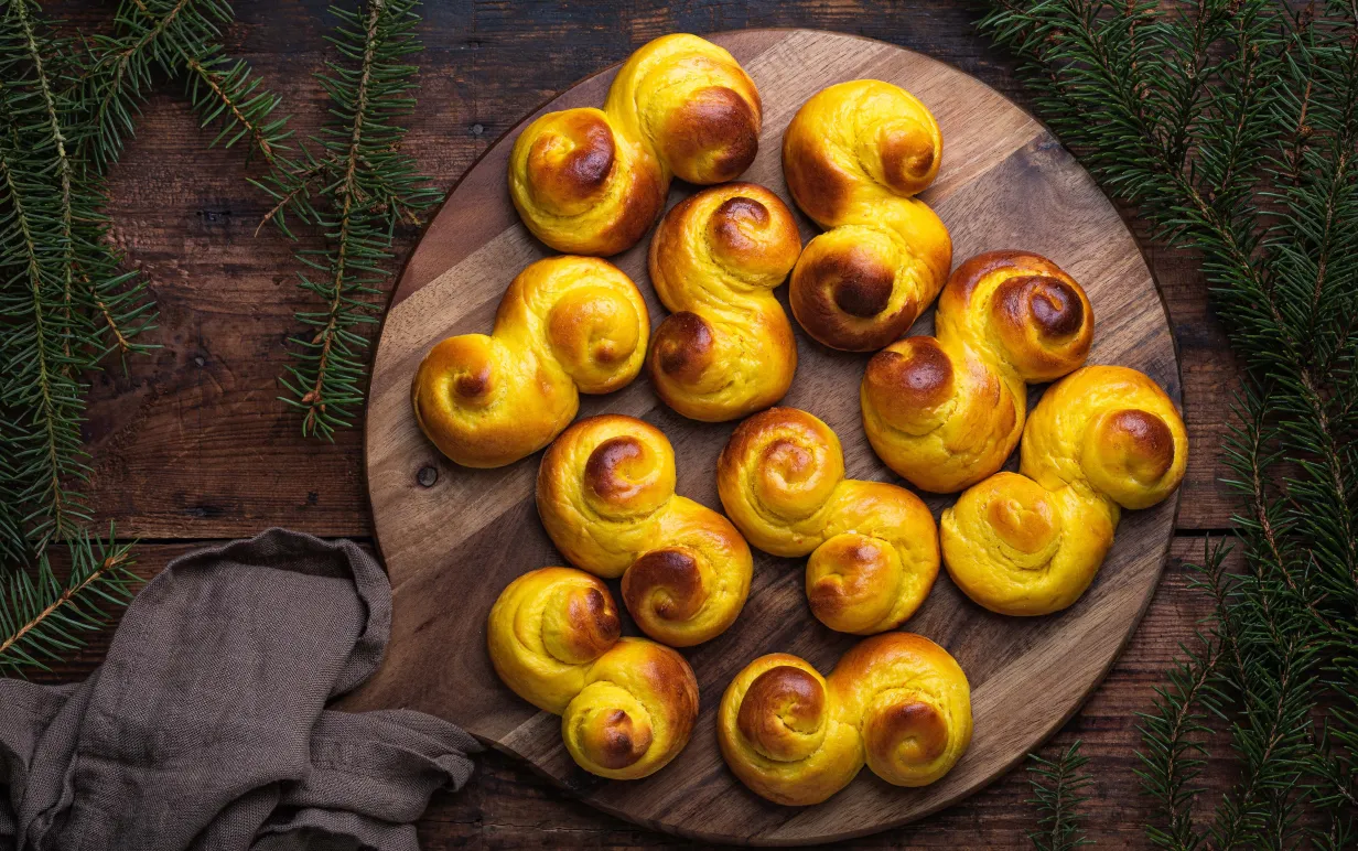 Freshly baked homemade Swedish traditional s-shaped saffron buns, also known as lussebullar or saffransbröd. Seen from above flat lay on a dark wooden table surrounded by spruce branches.