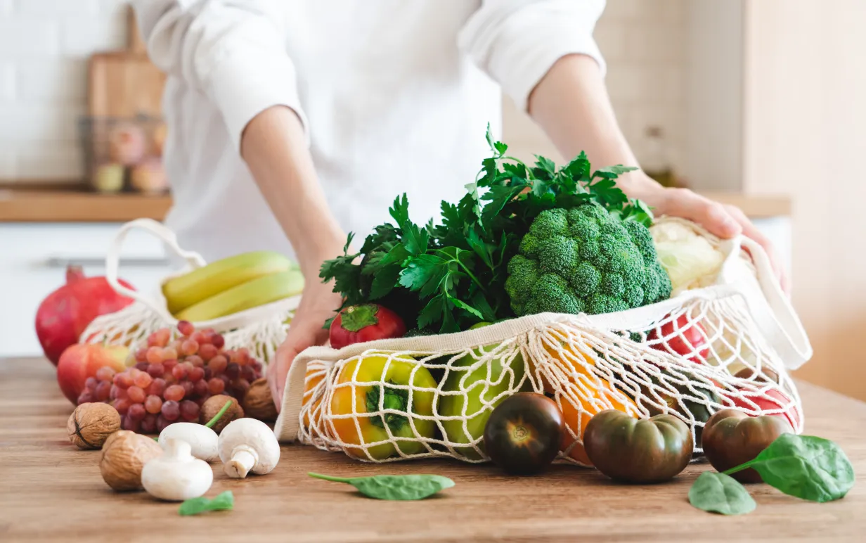 Fruits and vegetables in cotton reusable net mesh bag on wooden table in modern bright kitchen. Organic vegan food set. Sustainable lifestyle concept