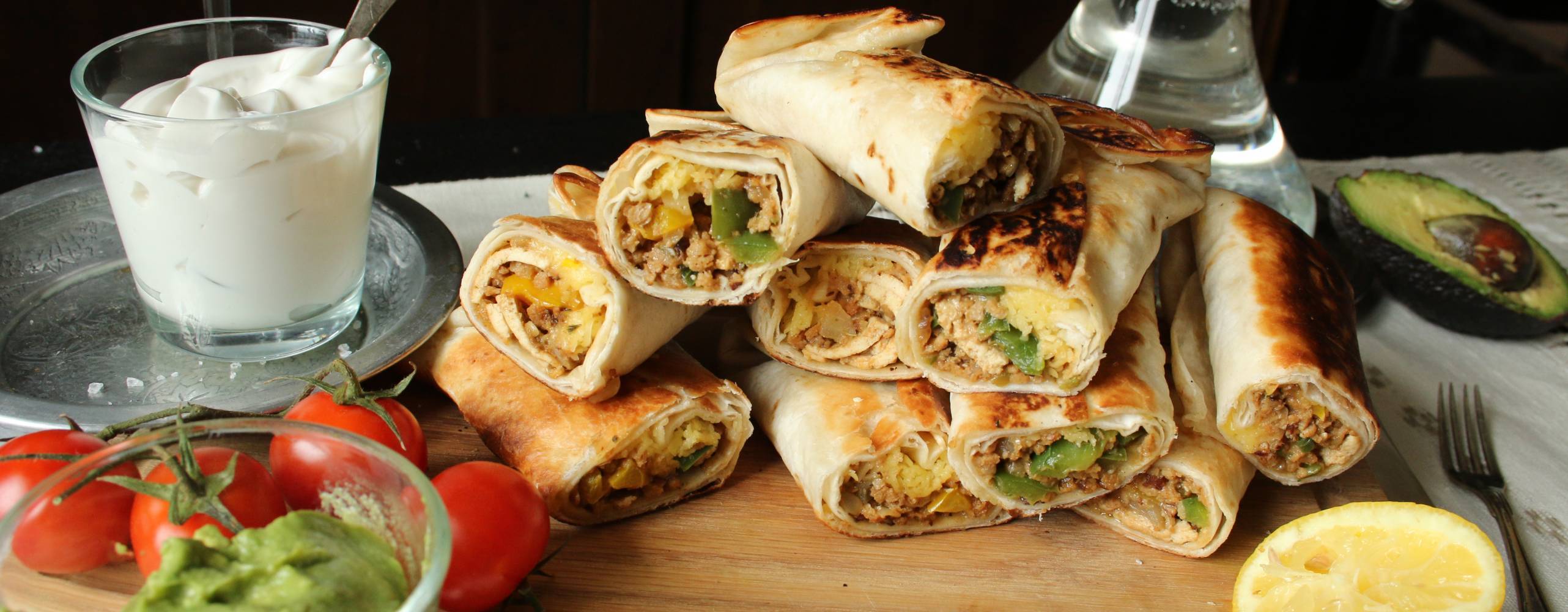 Chimichangas i en hög på en skärbräda, bredvid står en skål med vegansk créme fraiche och en skål med guacamole