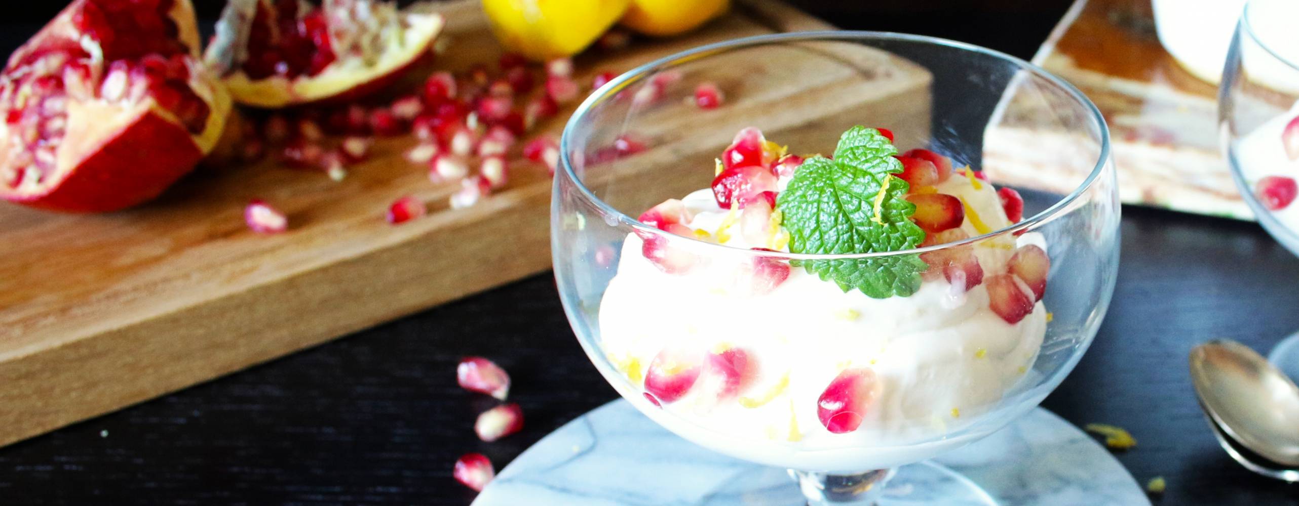 Glasskål med citronmousse och granatäppelkärnor framför en träskiva med granatäpplen och citroner.