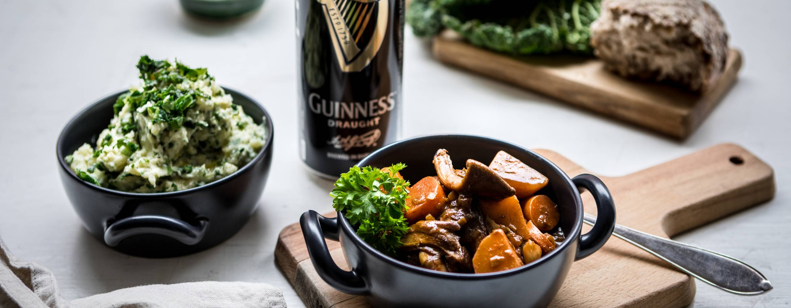 En skål med Irish stew står på en skärbräda bredvid en skål med irländskt potatismos och en öl