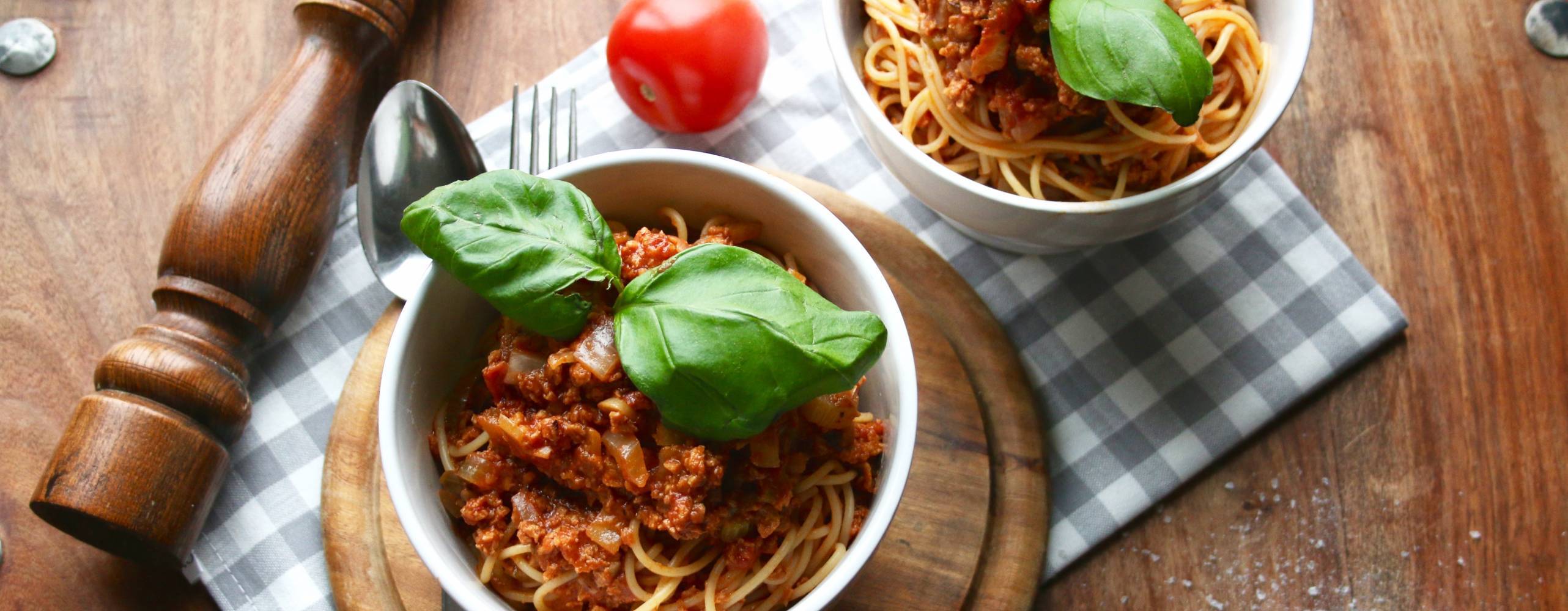 Två skålar med pasta bolognese på ett träbord. Det ligger en pepparkvarn och en tomat brevid skålarna.